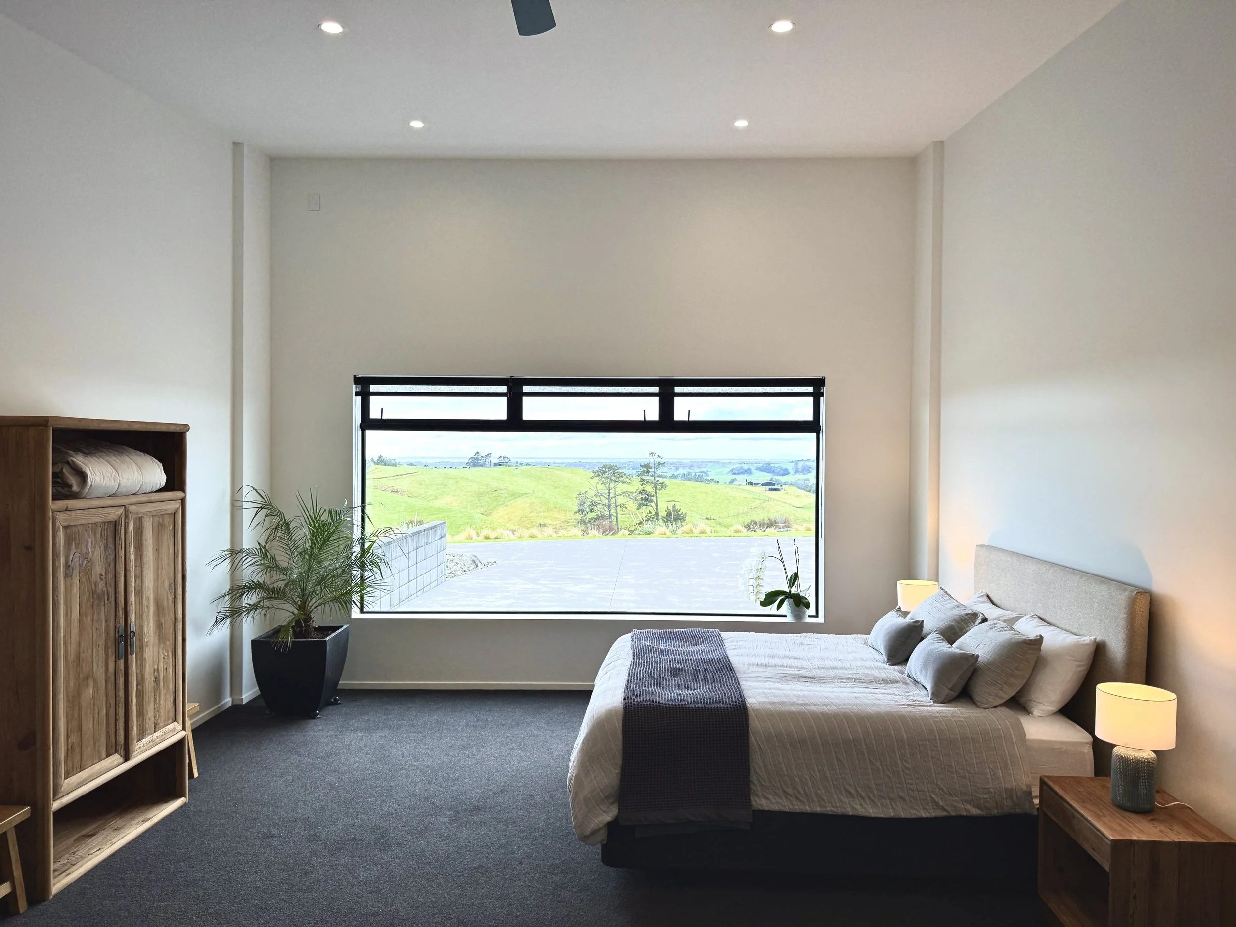 Modern bedroom with a neatly made bed, wooden wardrobe, potted plants, and a large window overlooking green rolling hills.