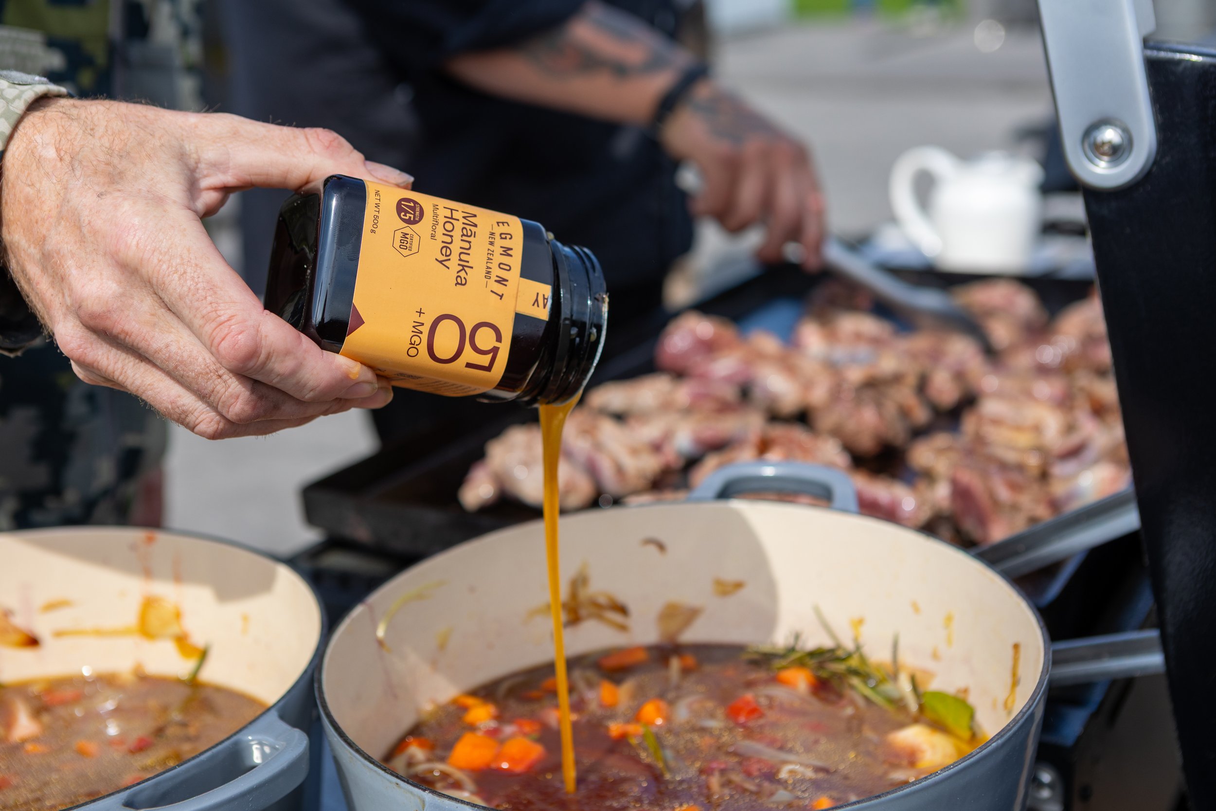 Person pours Manuka honey from a bottle into a pot of simmering stew outdoors.