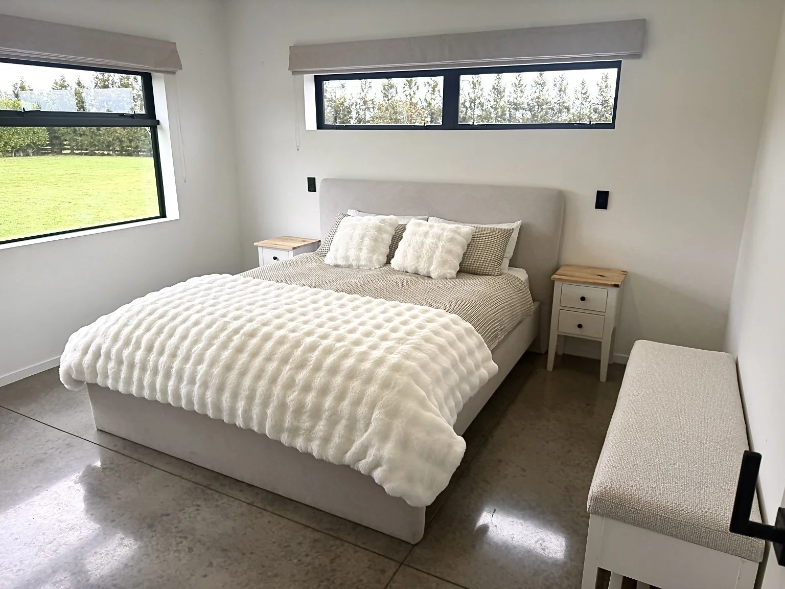 Bright modern bedroom with a white upholstered bed, textured duvet, bedside tables, large windows, and a cushioned bench.