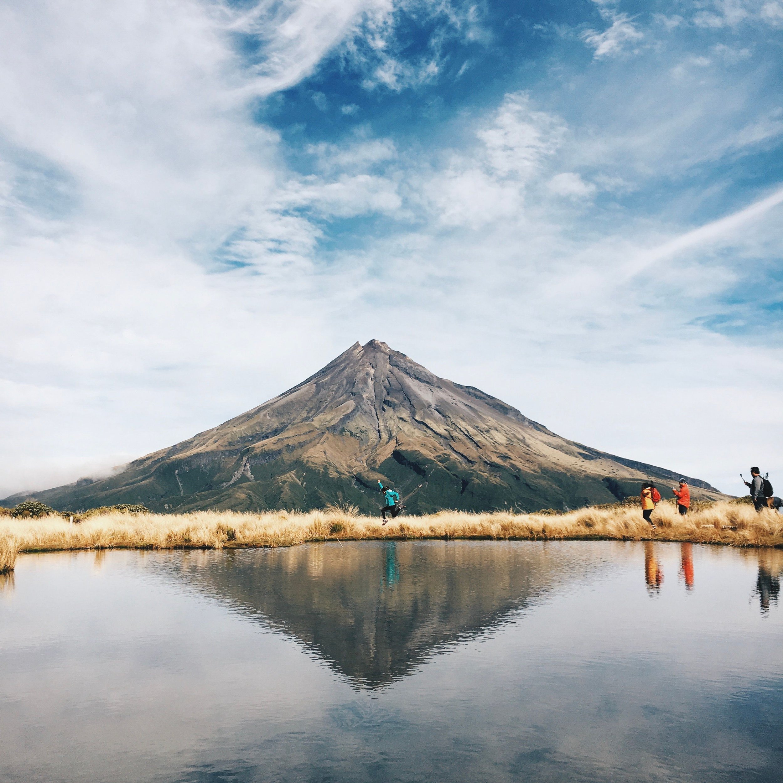 Hikers beside a reflective lake with a rugged volcanic mountain under a cloudy blue sky