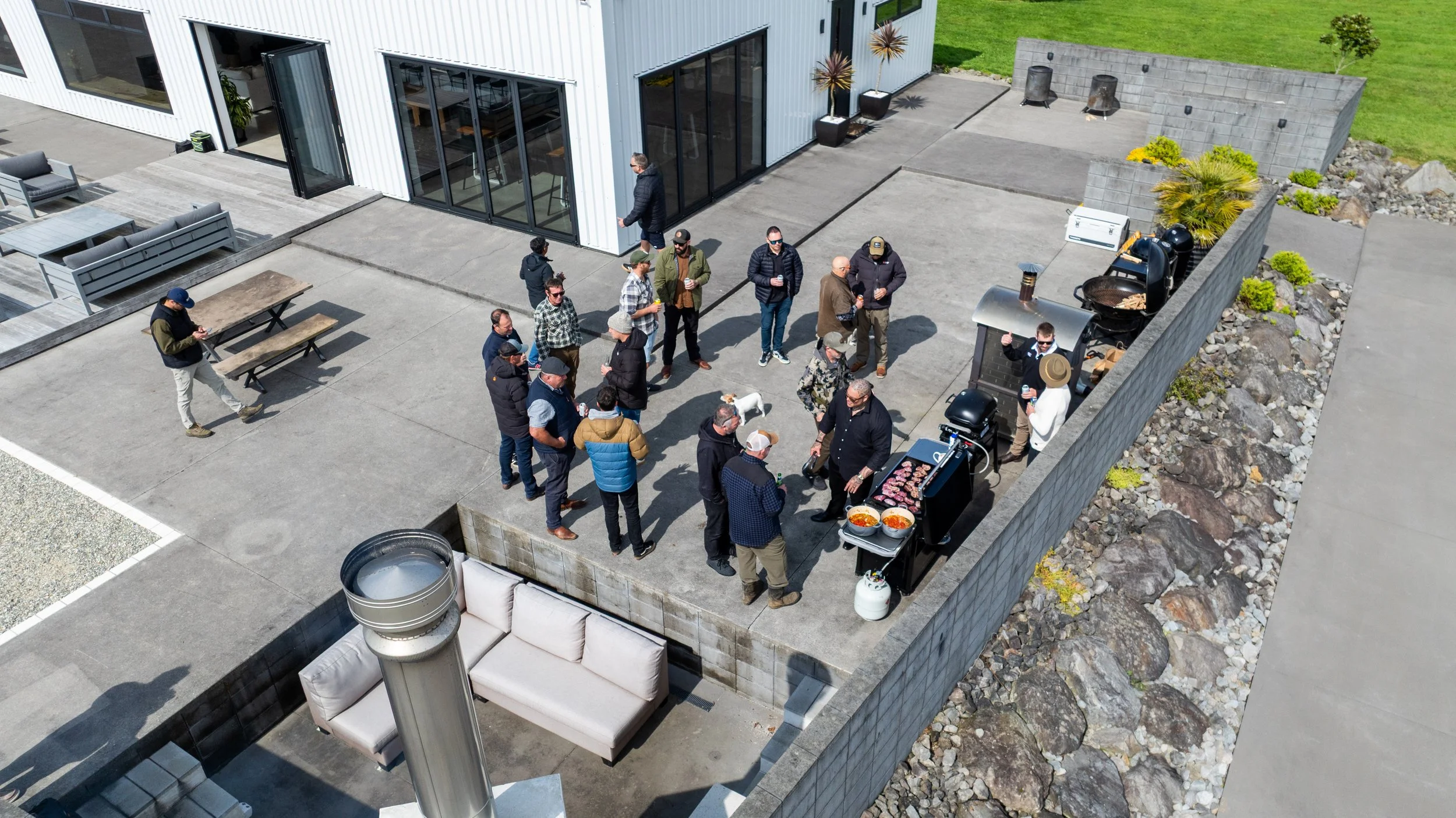 Aerial view of a group gathering on a modern patio for an outdoor barbecue, with grills, food, picnic tables, fire pits, and landscaped rock beds.