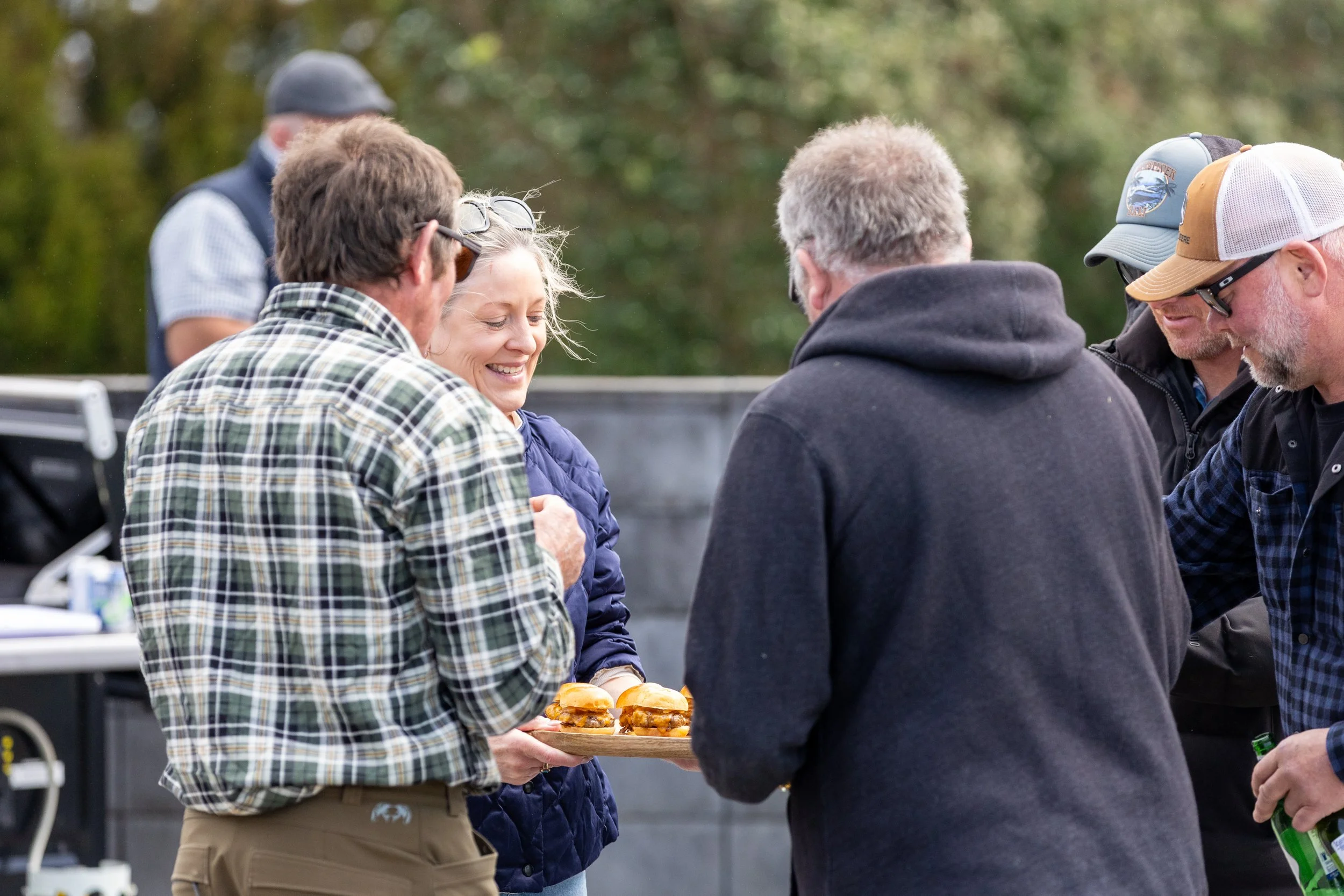 Group of adults socializing outdoors while a woman serves sliders on a wooden platter.