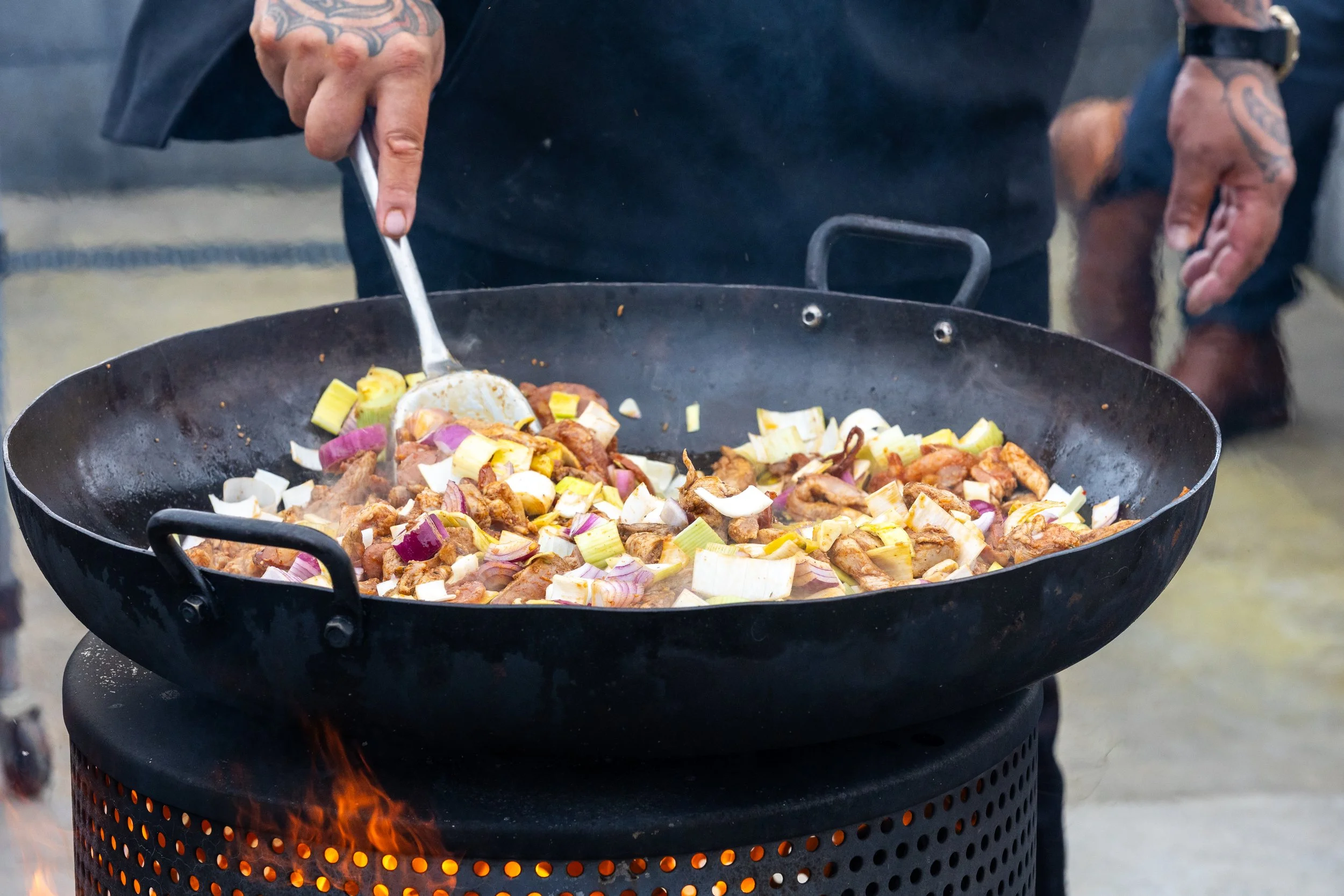 The Fat of the Land host stirring seasoned meat and chopped vegetables in a large outdoor skillet over an open flame
