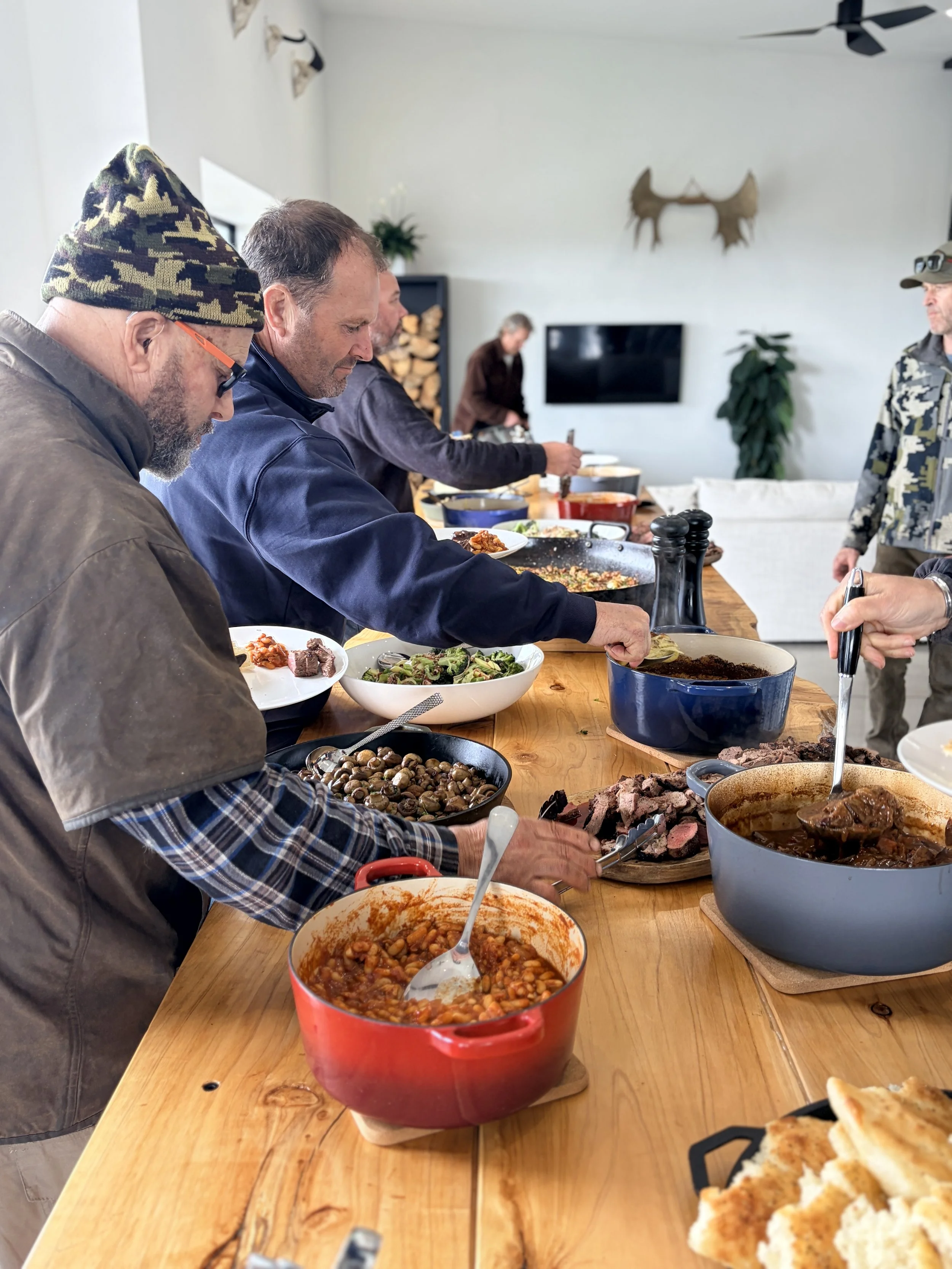 People sharing a buffet-style meal with roast beef, vegetables, mushrooms, beans, and bread around a wooden dining table.