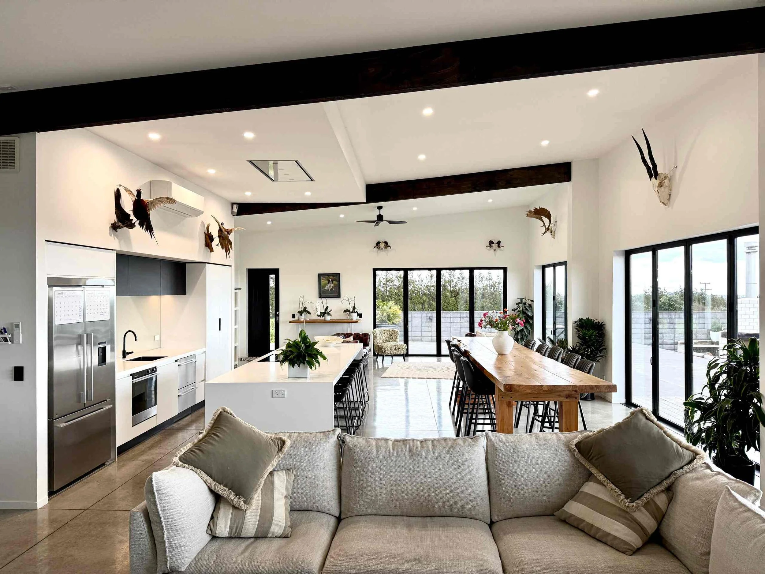 Modern open-plan home interior with kitchen, island, dining table, living room sofa, plants, large windows, and mounted bird and animal decor