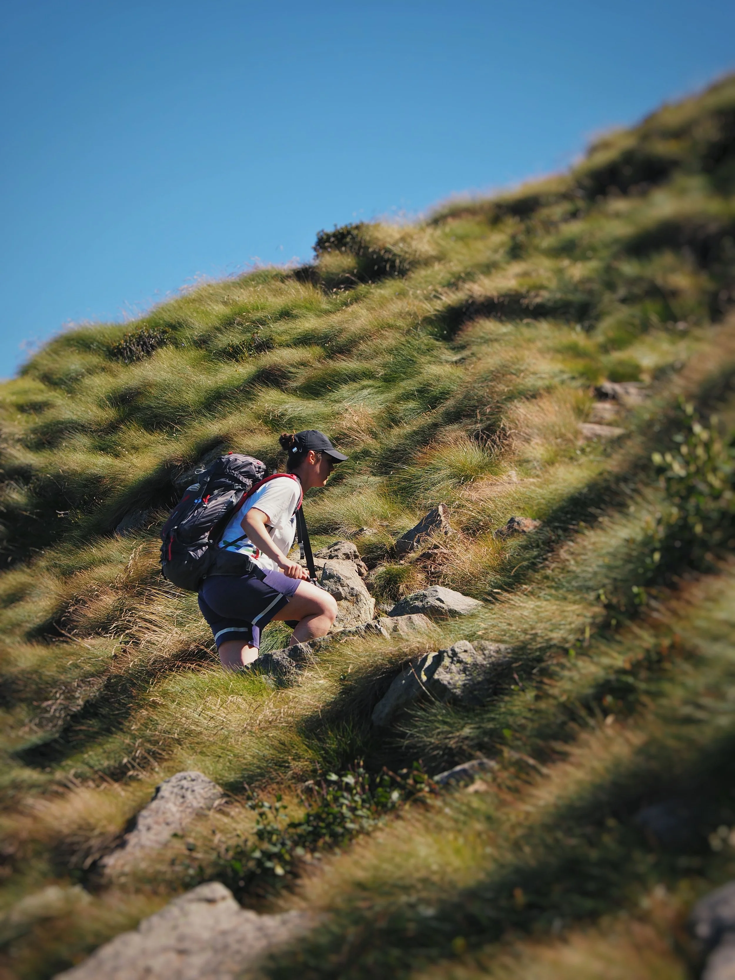 Hiker with a backpack climbing a rocky, grassy mountain trail under a clear blue sky
