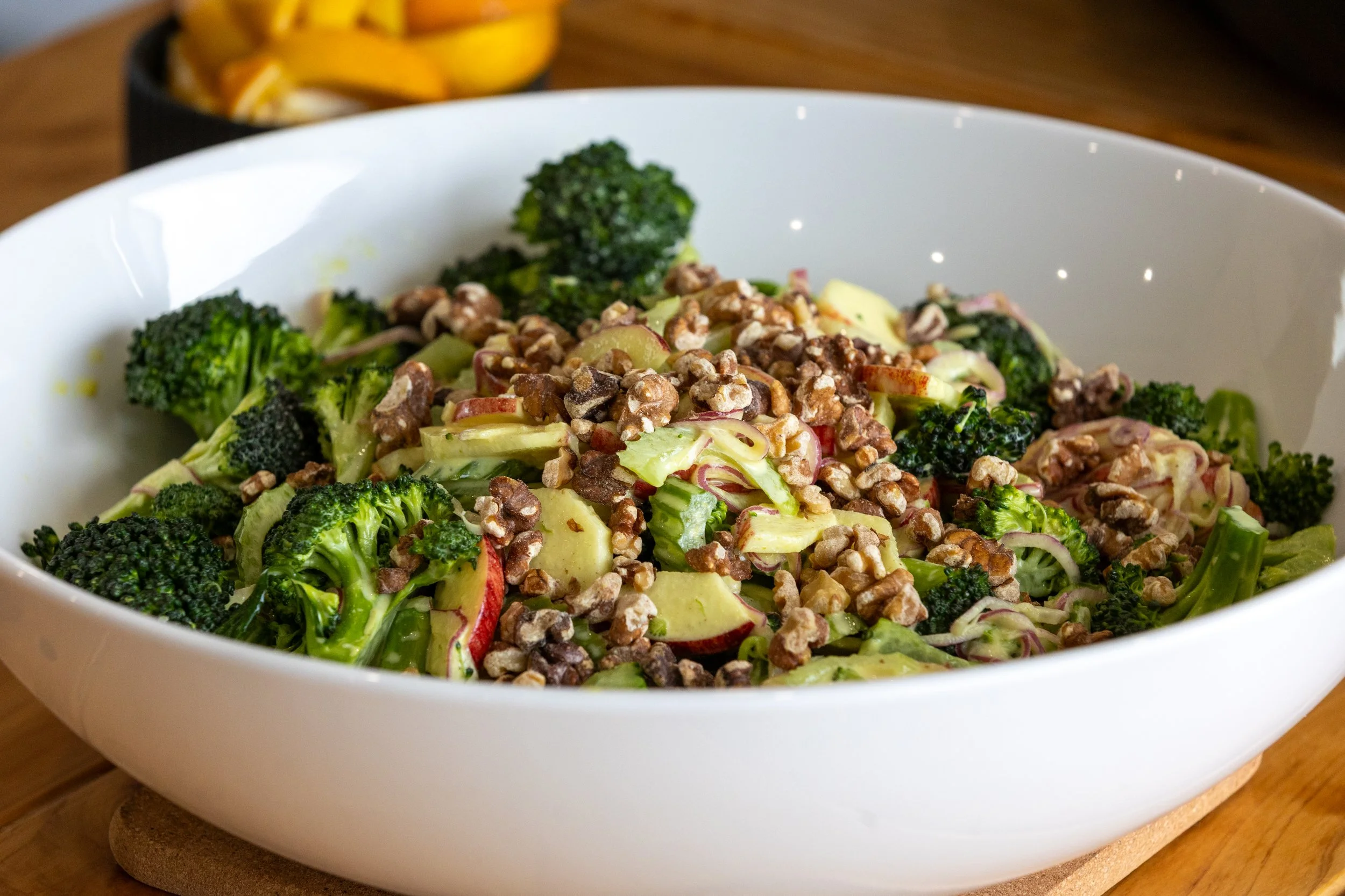 Broccoli salad with sliced apples, red onion, creamy dressing, and chopped walnuts in a white serving bowl
