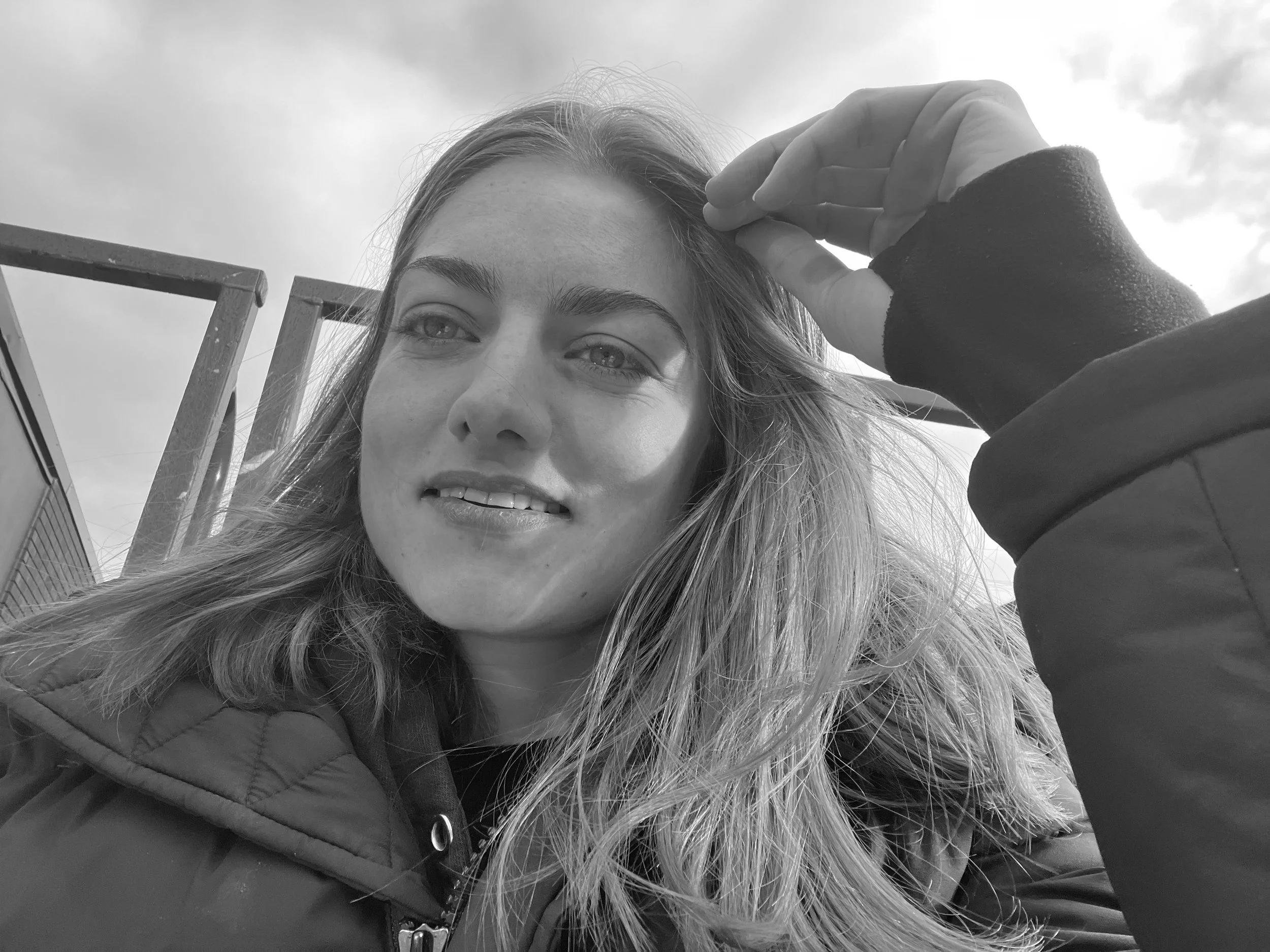 A black and white photo of a young woman with light-colored hair, smiling slightly, with her hand partially covering her forehead. She is wearing a dark jacket and standing outdoors with an overcast sky in the background.