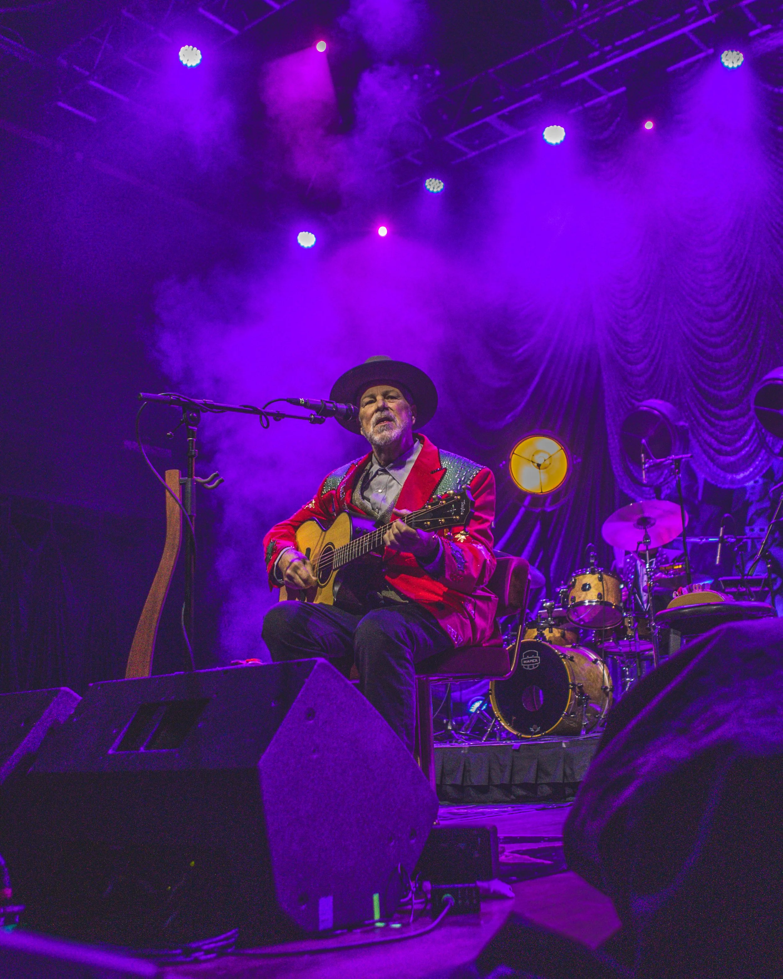 A musician with a beard, wearing a wide-brimmed hat and a red and green jacket, is playing an acoustic guitar on stage. The stage is lit with purple and blue lights, with a drum set in the background and purple velvet curtains.