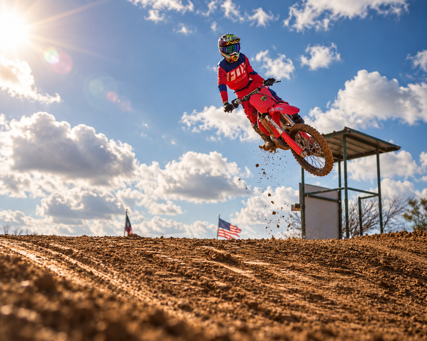 A person riding a red dirt bike jumps off a ramp on a dirt track during daytime, with the sun shining and clouds in the sky, and American flags in the background.