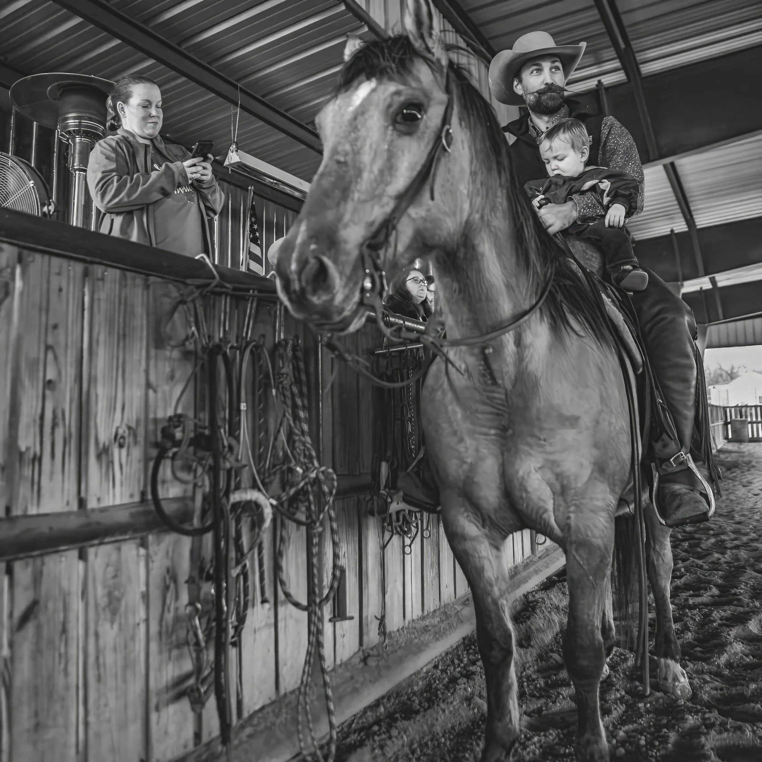 A black and white photo of a horse with a rider wearing a cowboy hat, standing inside a barn with a woman on the left using her phone and another woman on the right holding a child.
