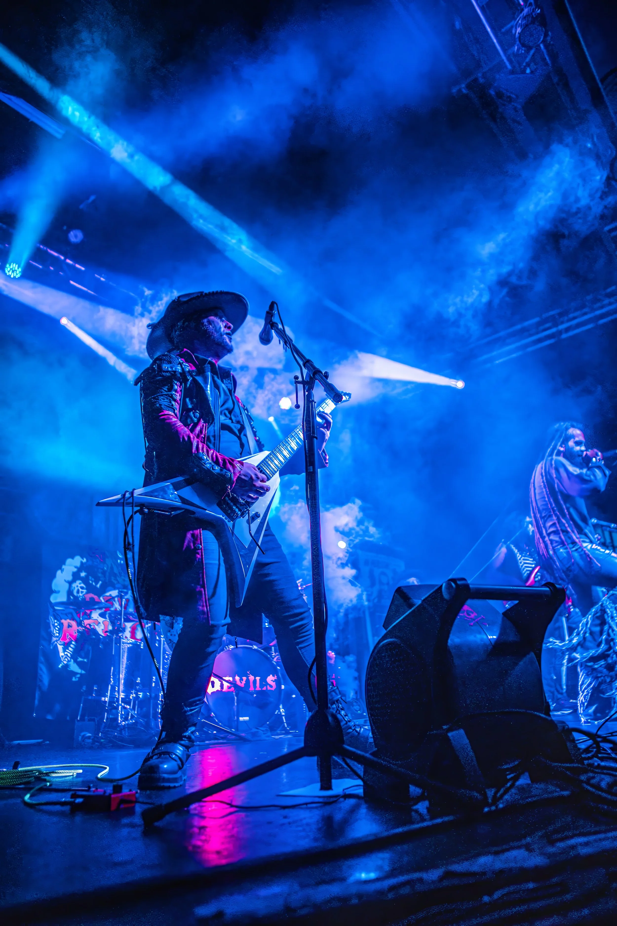 A musician playing an electric guitar on stage amidst blue and purple lights with smoke effects, wearing a cowboy hat and artistic clothing.