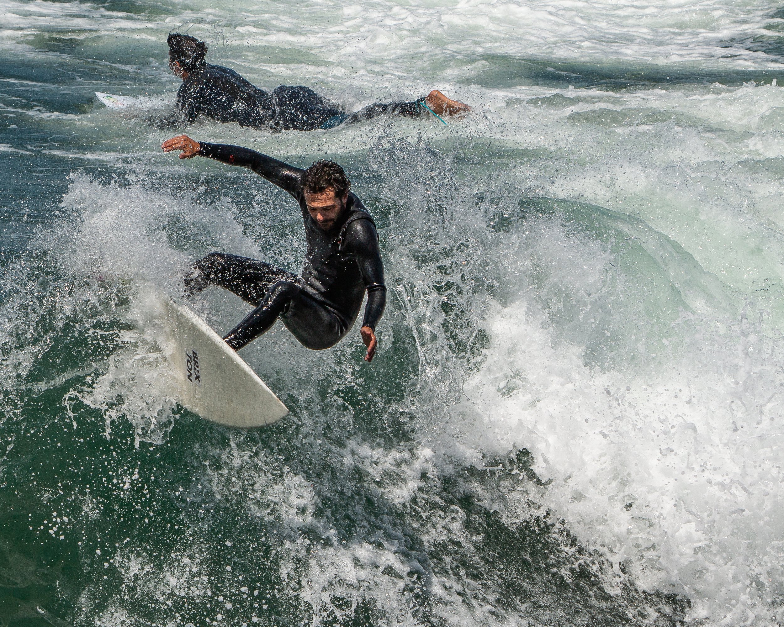 A man in a wetsuit is surfing on a wave in the ocean, with water splashing around him and another person behind him also riding the wave.