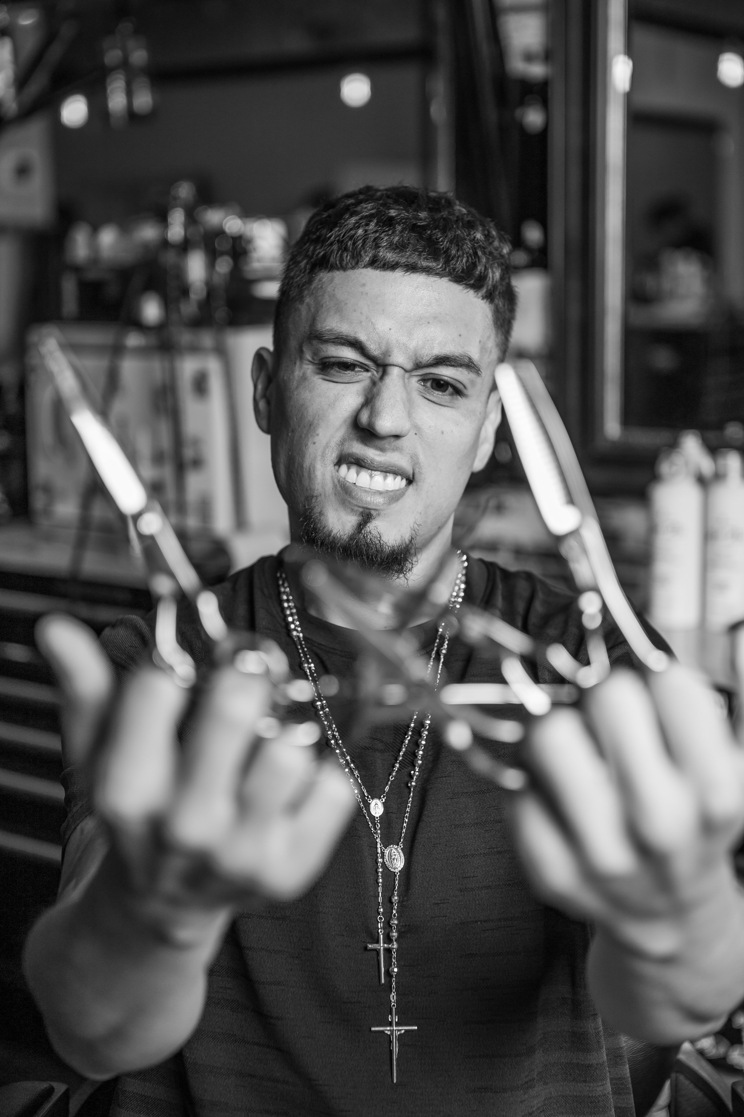 Black and white photo of a man with short curly hair and goatee, smiling with one eye closed, reaching towards the camera with both hands, wearing a dark shirt and multiple necklaces, standing in a room with shelves and various objects behind him.