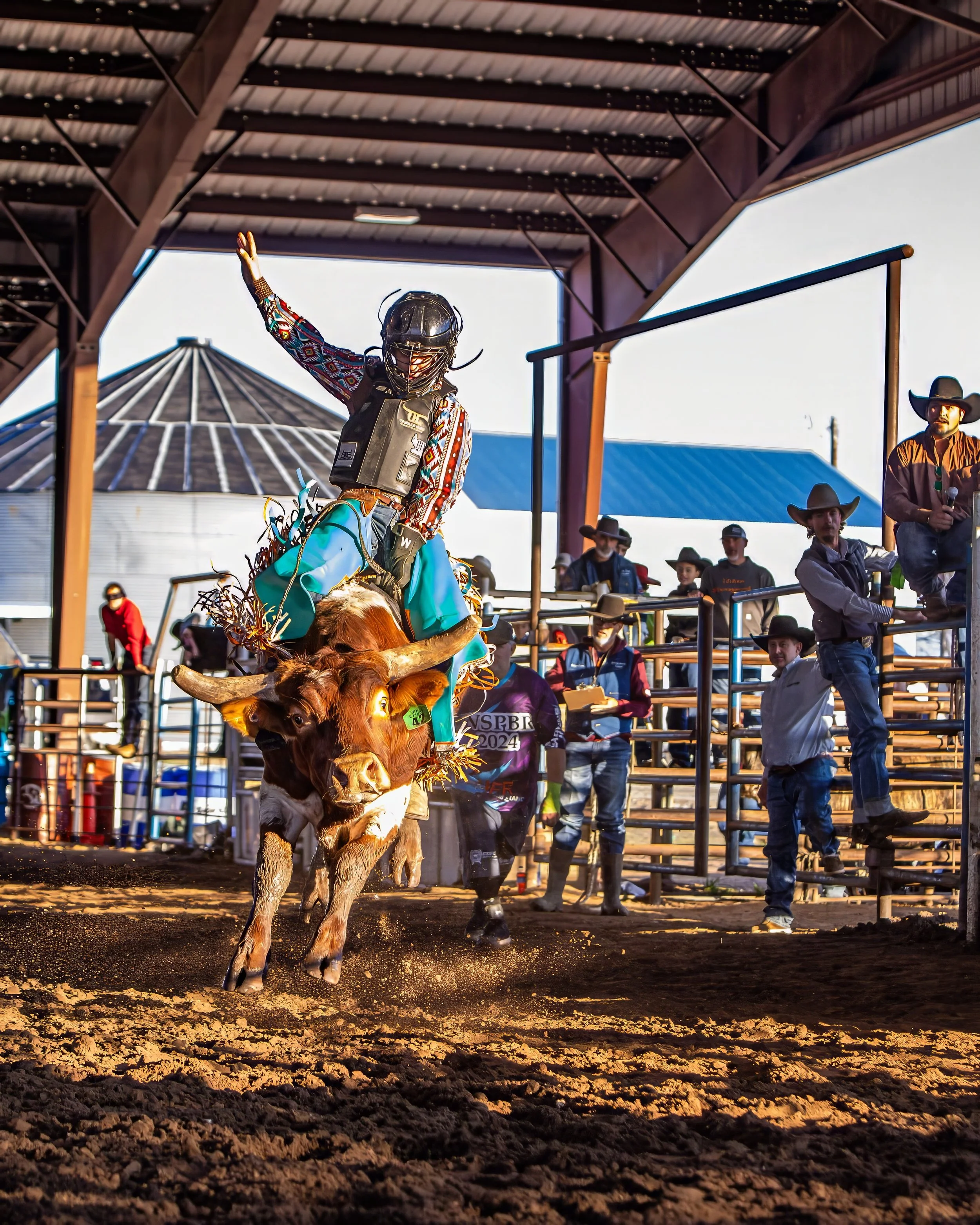 A cowboy riding a bucking bull during a rodeo event inside a large covered arena, with spectators watching from the stands.