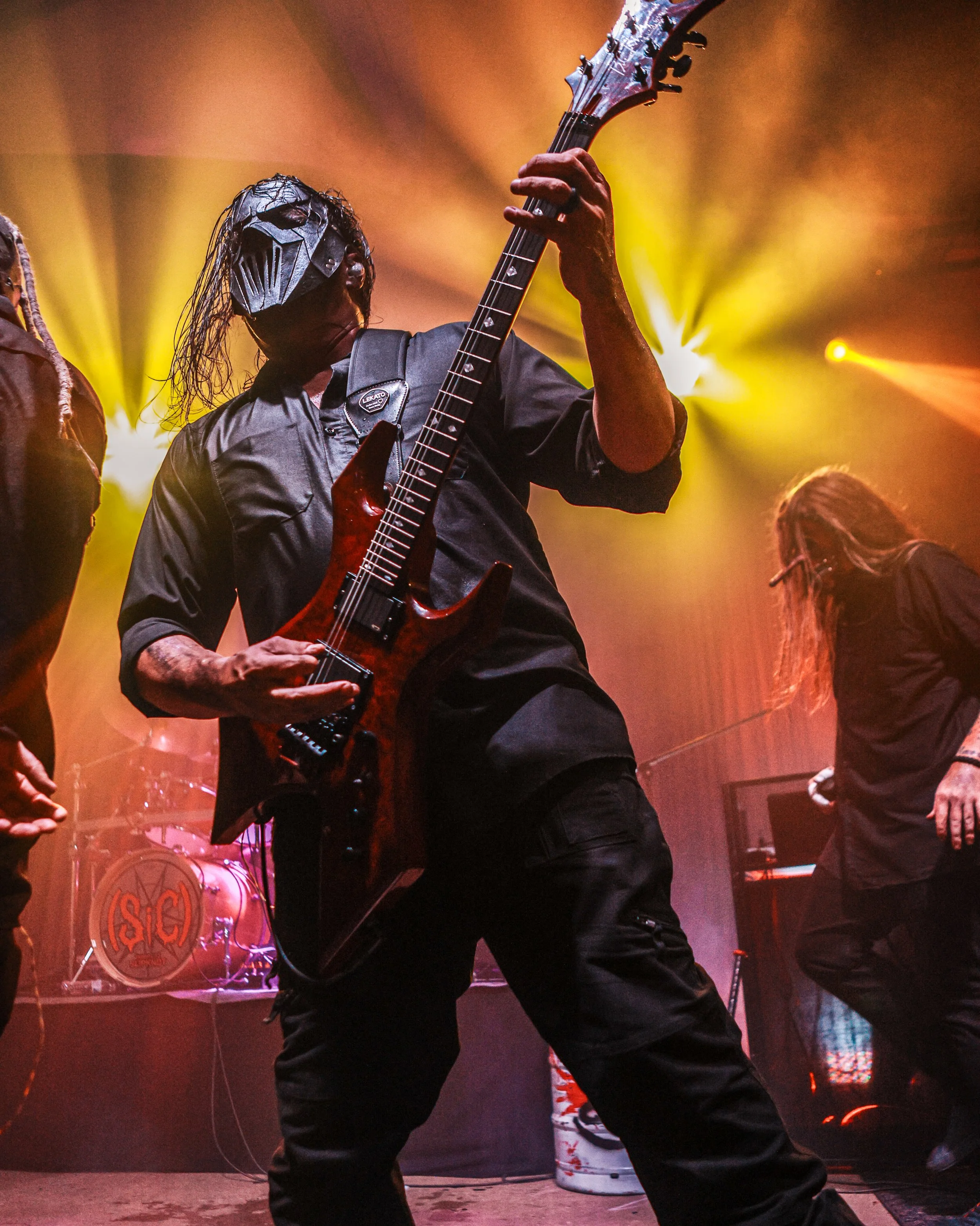 A musician wearing a dark helmet and black shirt plays an electric guitar on stage with colorful concert lighting.