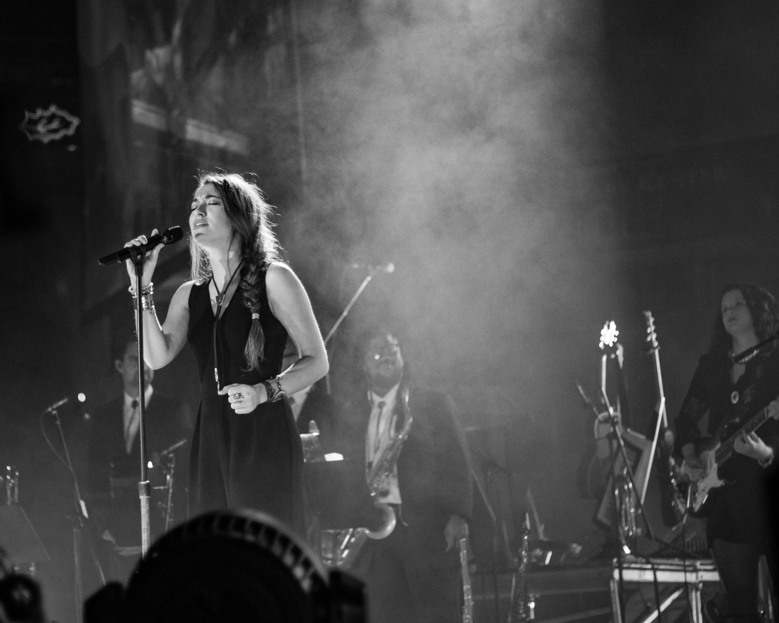 A woman singing into a microphone on stage with eyes closed, surrounded by stage lights and musical equipment, in black and white.