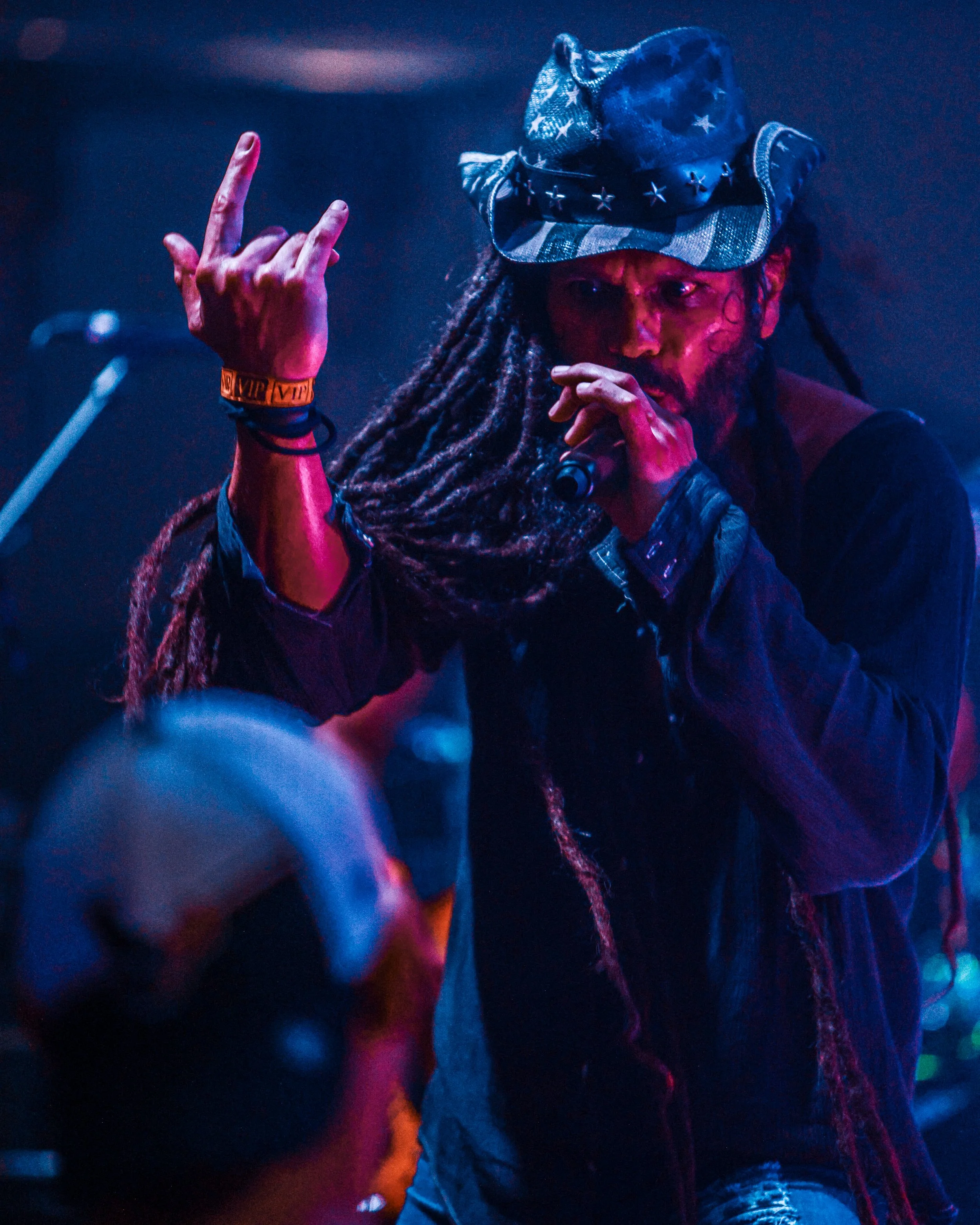 A person with dreadlocks, wearing a camouflage hat with metal stars, performing on stage with colorful lights, holding a microphone, and making a hand gesture.