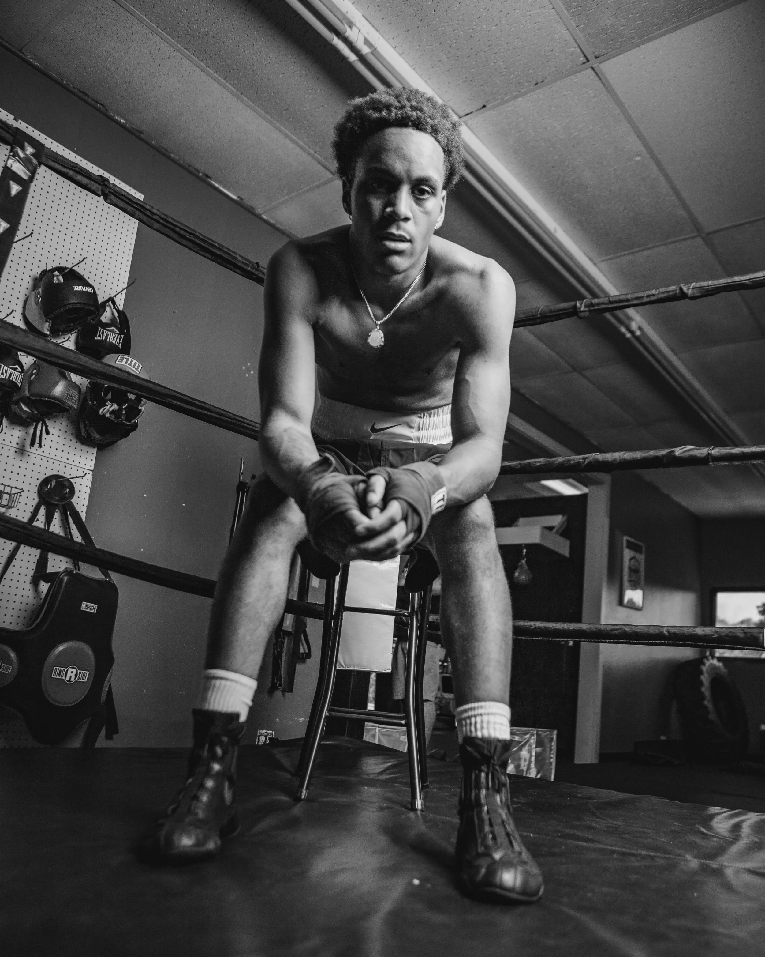 Black and white photo of a young male boxer in a gym, leaning forward with hands clasped, wearing boxing gloves, shorts, and a necklace, surrounded by boxing equipment and rings.