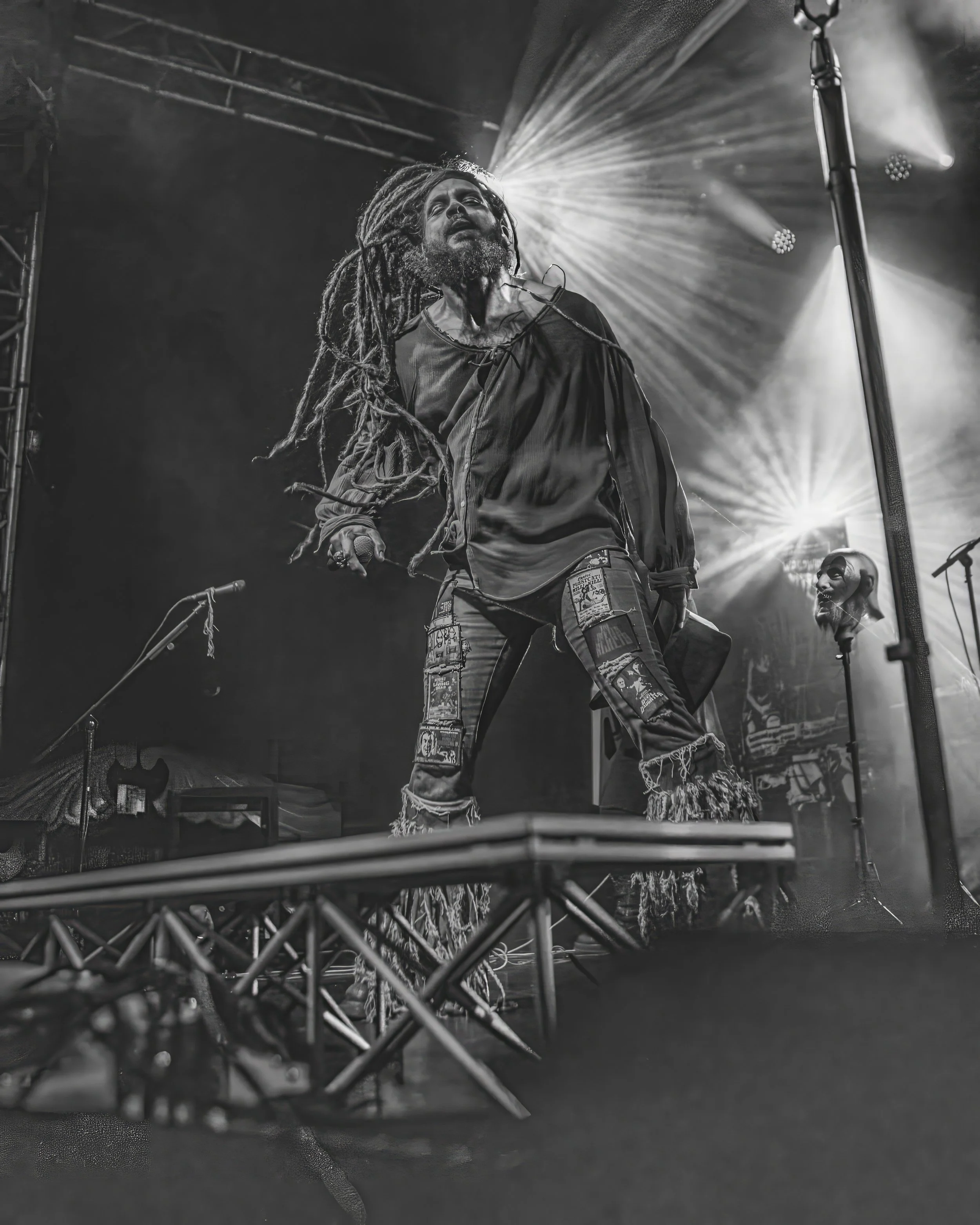 Black and white photo of a man with dreadlocks singing into a microphone on stage, with stage lights shining behind him, and a person with a shaved head and beard in the background.