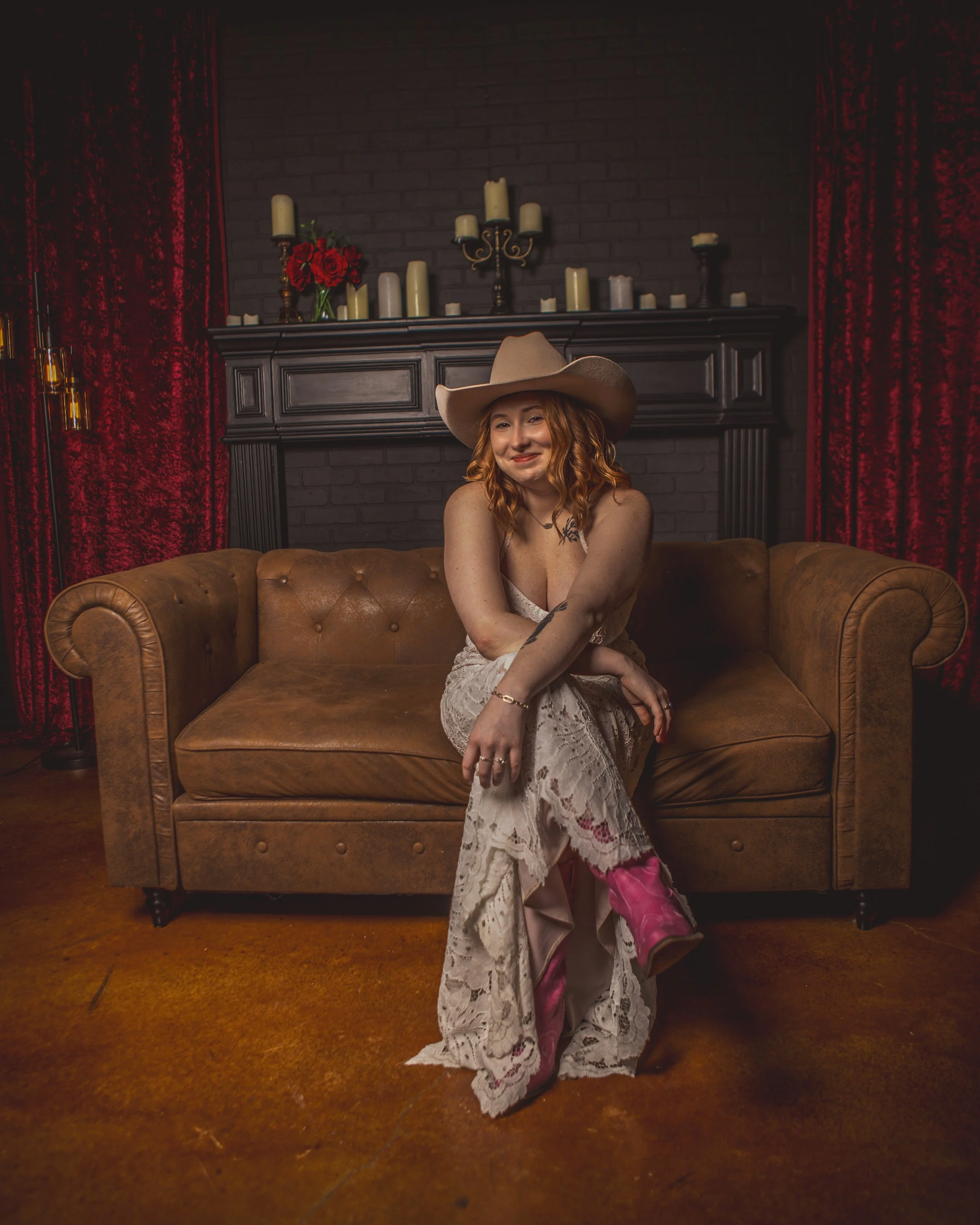 A woman with red hair sitting on a brown leather couch in front of a black brick fireplace, wearing a large cowboy hat, a lace dress with a tattoo on her left arm, and pink cowboy boots. The fireplace mantle is decorated with candles and red flowers.
