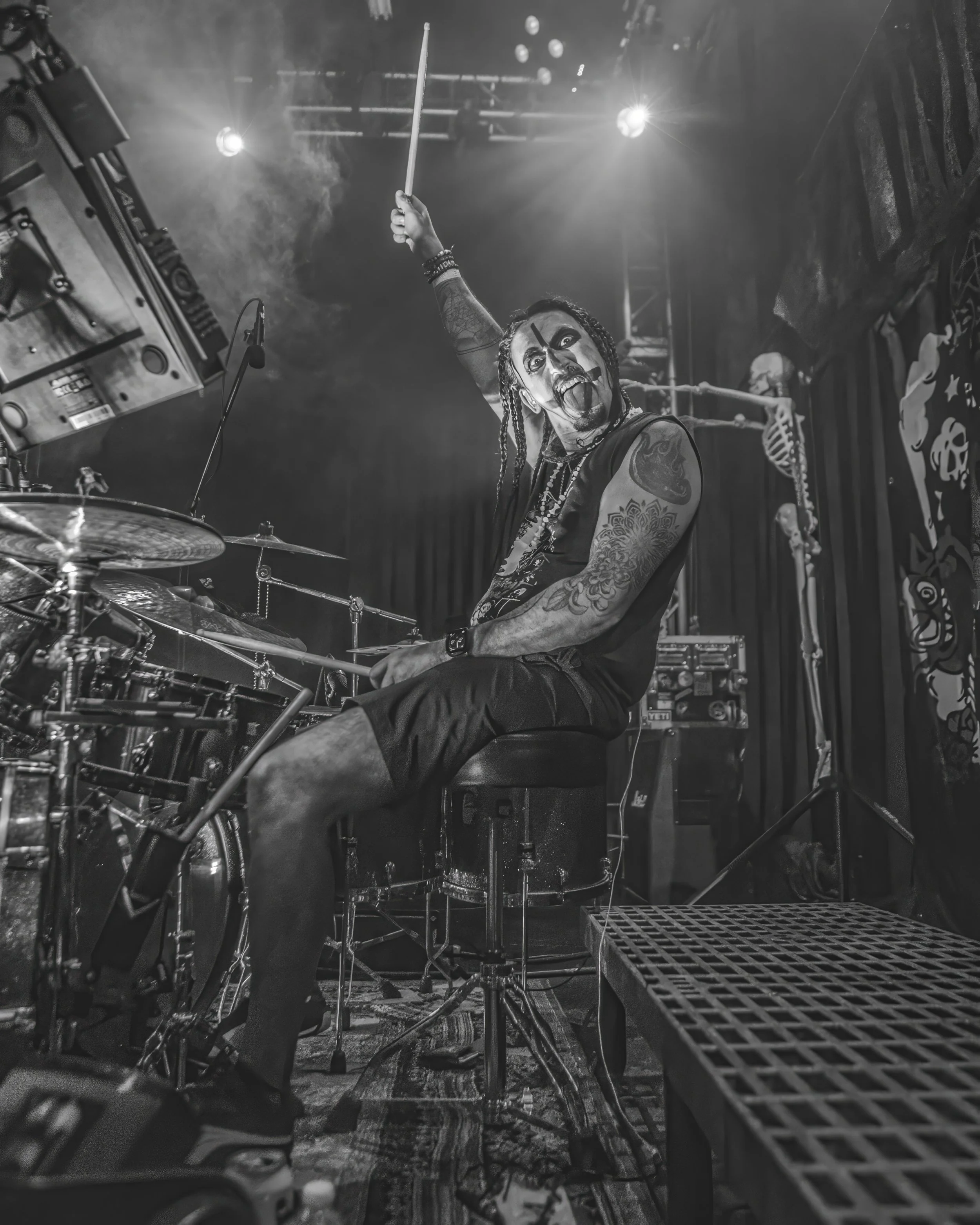 A drummer with tattoos, wearing makeup resembling a scary clown or Joker, raising a drumstick in a performance setting with stage lights and skeleton decorations in the background.