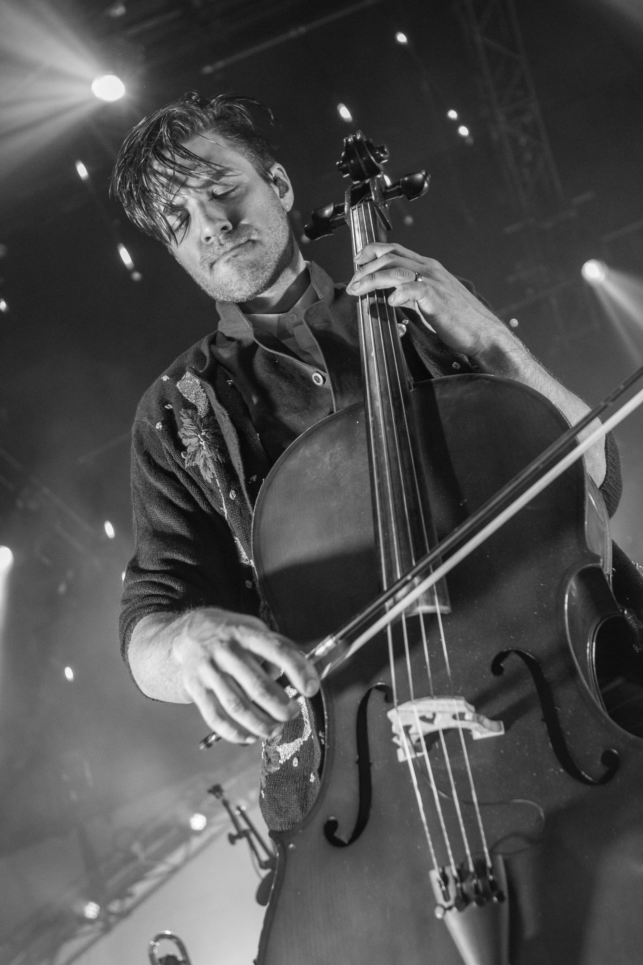 A man playing a cello on stage, with closed eyes and focused expression, illuminated by stage lights in a concert setting.