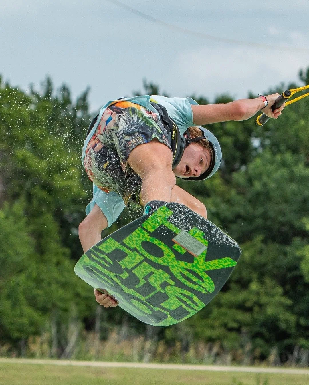 A male wakeboarder in mid-air performing a trick with a green and black wakeboard, wearing a helmet, a harness, and colorful shorts, with water splashing around him and a background of green trees.