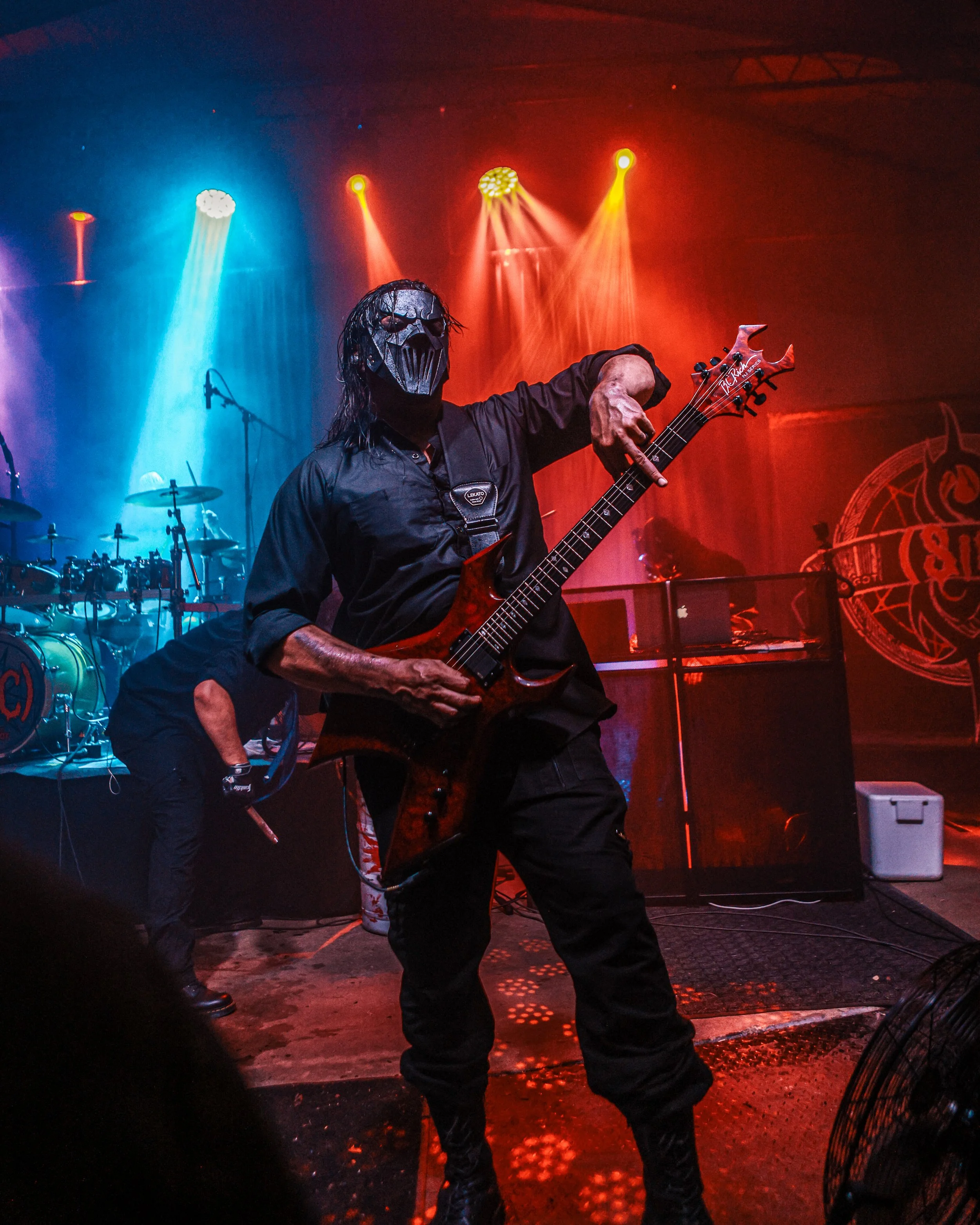 A person wearing a metallic skull mask and sunglasses playing an electric guitar on stage with colorful stage lights and equipment visible in the background.