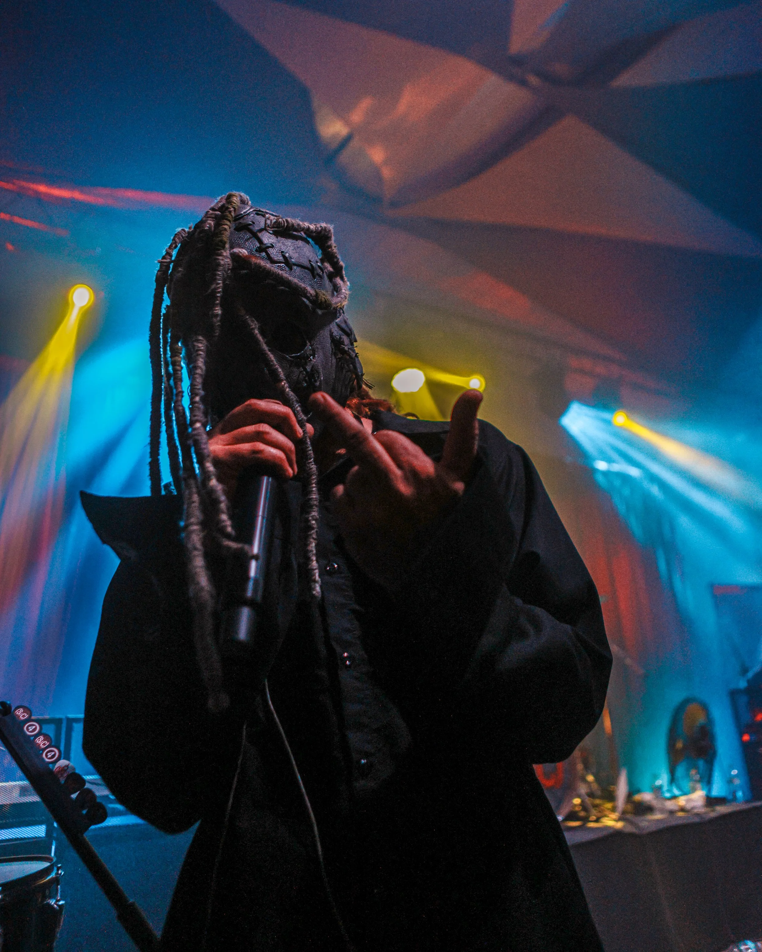Person with dreadlocks wearing a black face mask and black jacket, holding a microphone, standing in a colorful, illuminated stage or event setting.