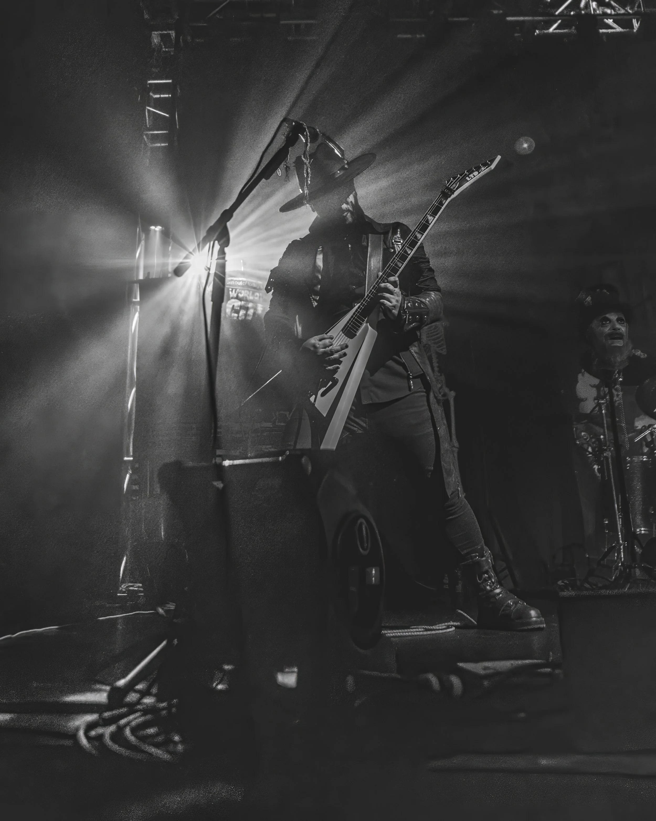 A black and white photo of a man wearing a cowboy hat and leather jacket, holding an electric guitar on stage with bright light rays behind him. To the right, another person is visible, smiling and looking upward.