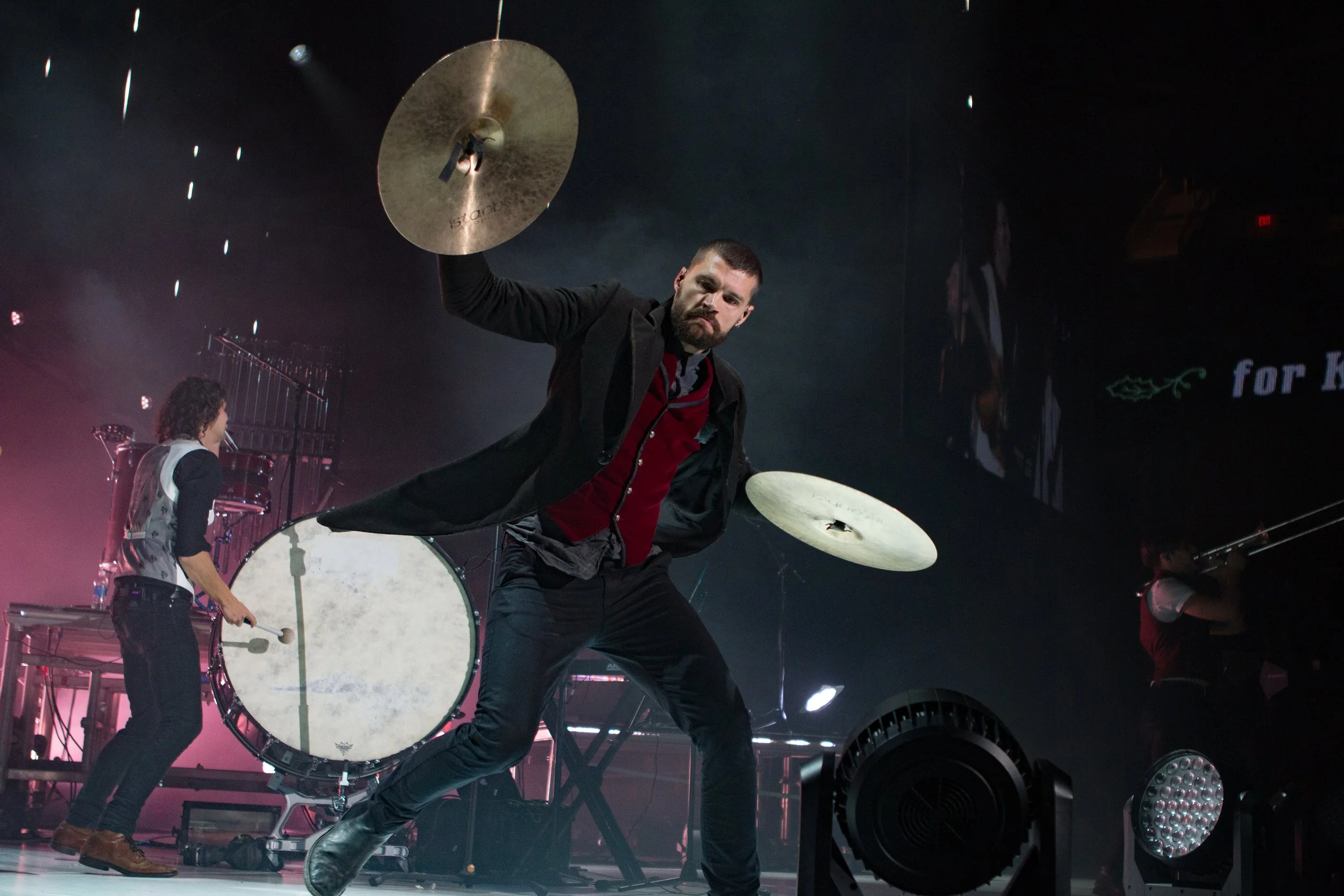 Performer playing drums on stage with a cymbal in the air. Concert lighting and other musicians are visible in the background.
