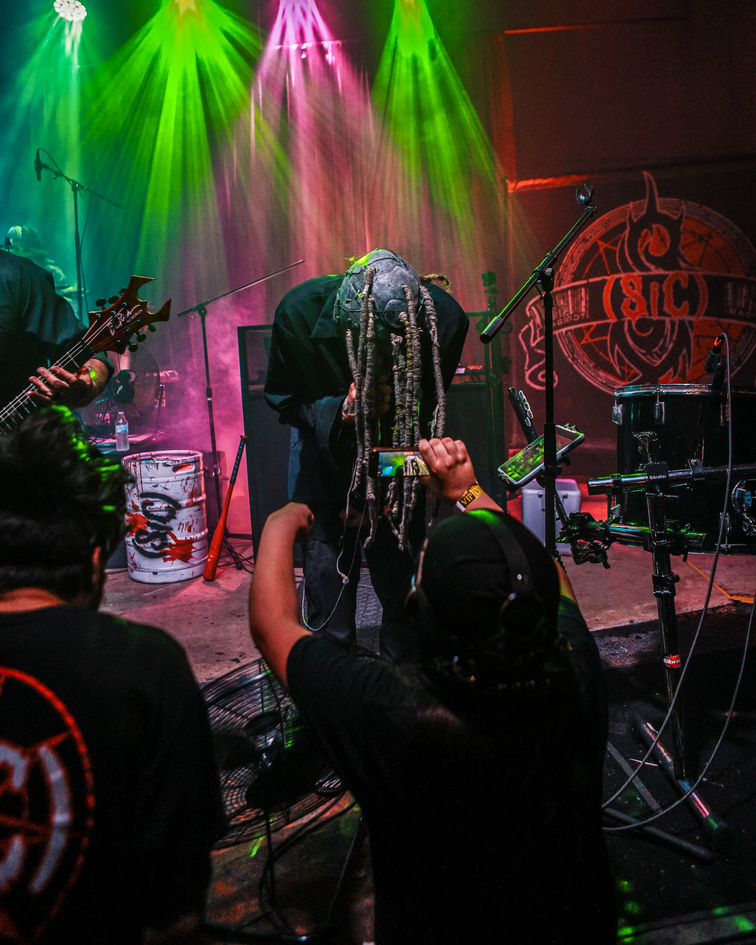 A musician with long dreadlocks on stage at a concert lit with green and pink stage lights. There are microphones, musical equipment, and audience members with raised hands in the foreground.
