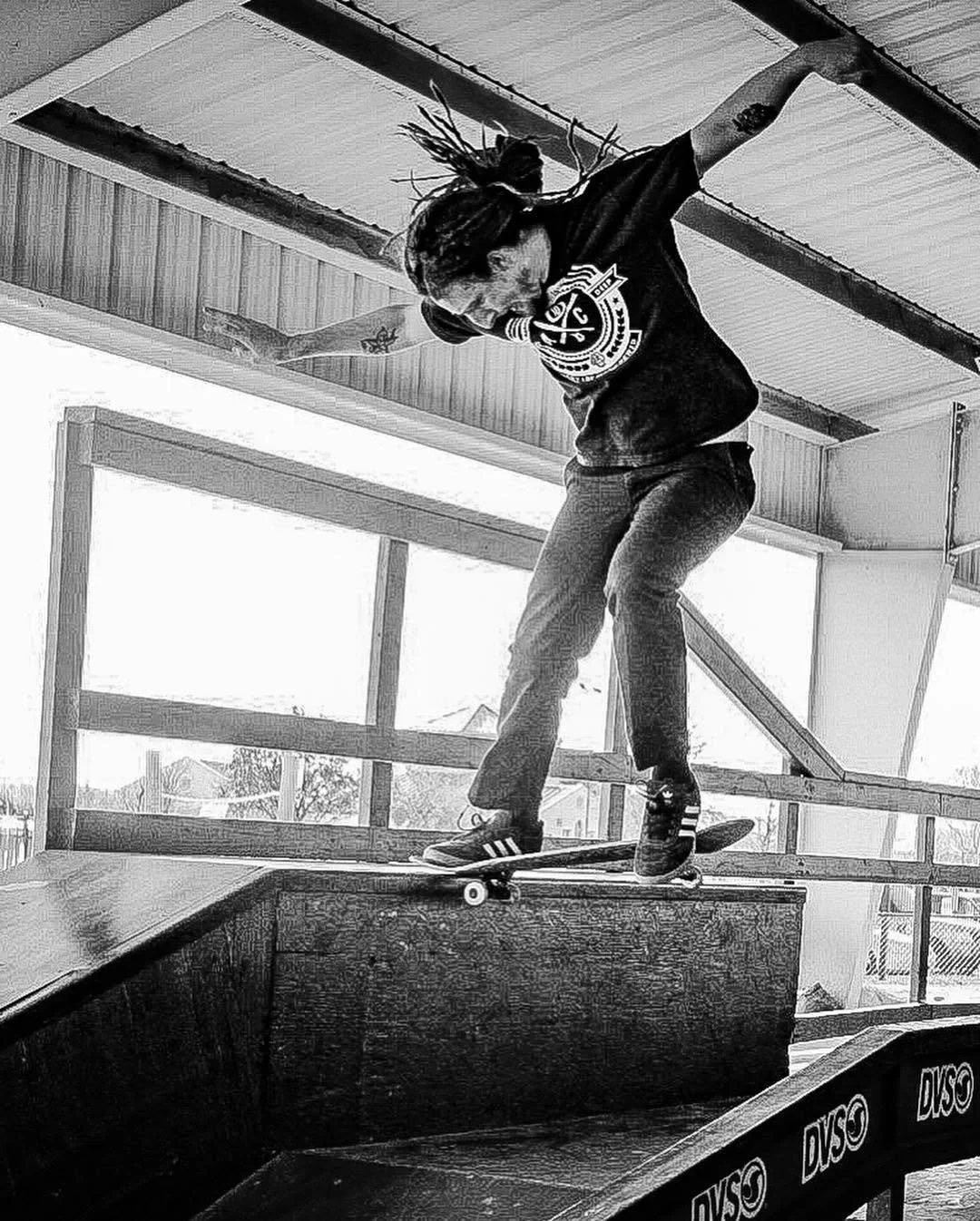 Skateboarder performing a trick on a skate ramp indoors, with wooden paneling and windows in the background.