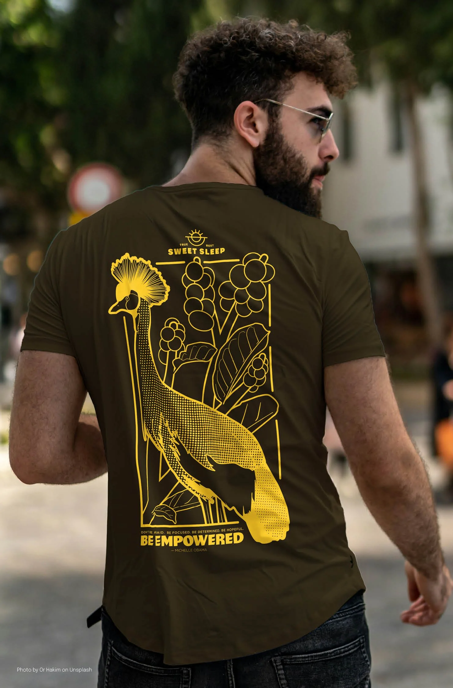 Man with sunglasses modeling the Uganda themed "Sweet Sleep" outside. It's a dark brown shirt with a gold ink graphic of a Grey Crowned Crane, a coffee plant, the Sweet Sleep logo at the top and a quote by Michelle Obama at the bottom.