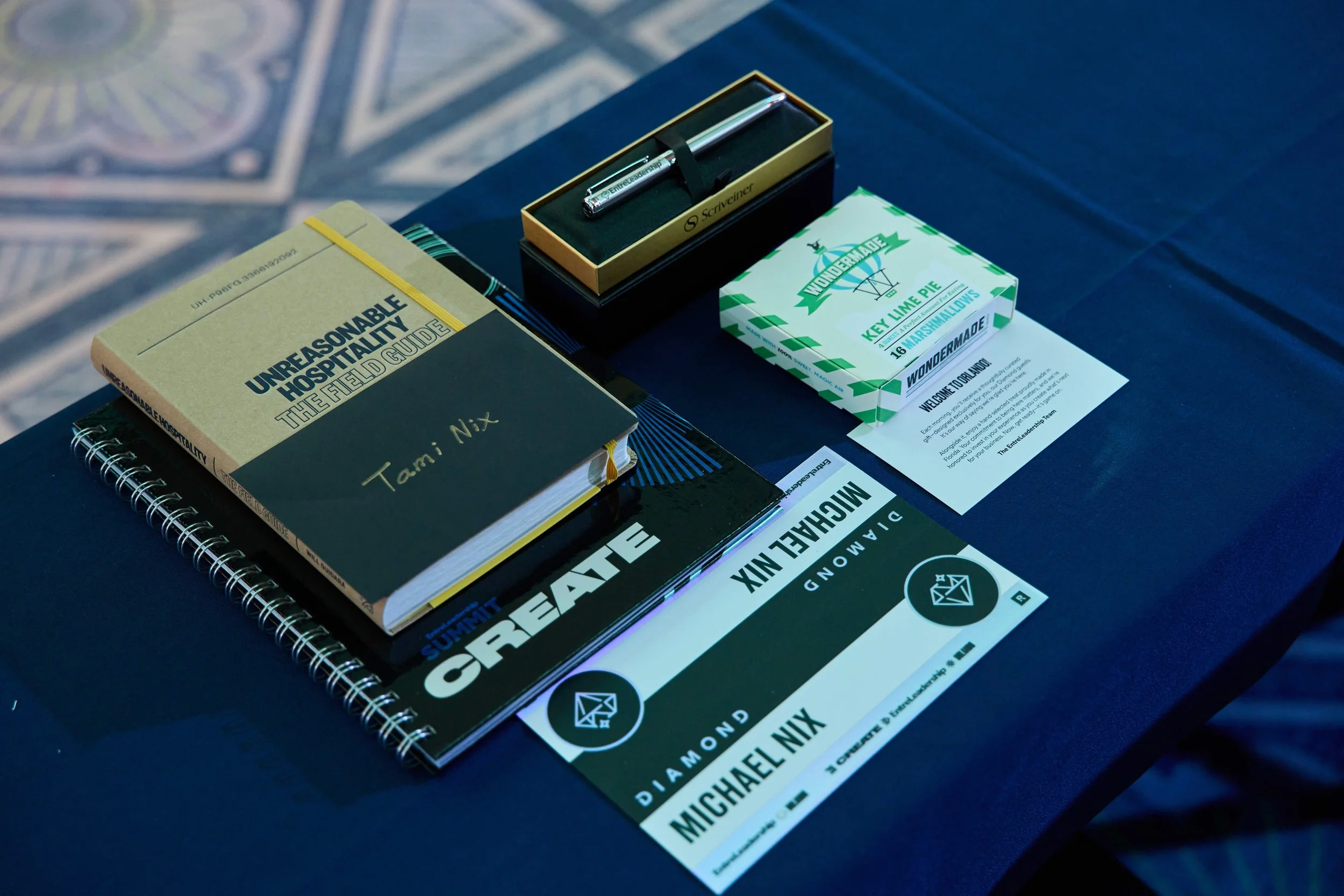 Gifts that Diamond tier attendees get at their seat. It includes the Summit Create workbook, a Diamond name tent, a welcome card, branded pen, Will Guidara's book, a personalized note, branded fountain pen, and a snack.