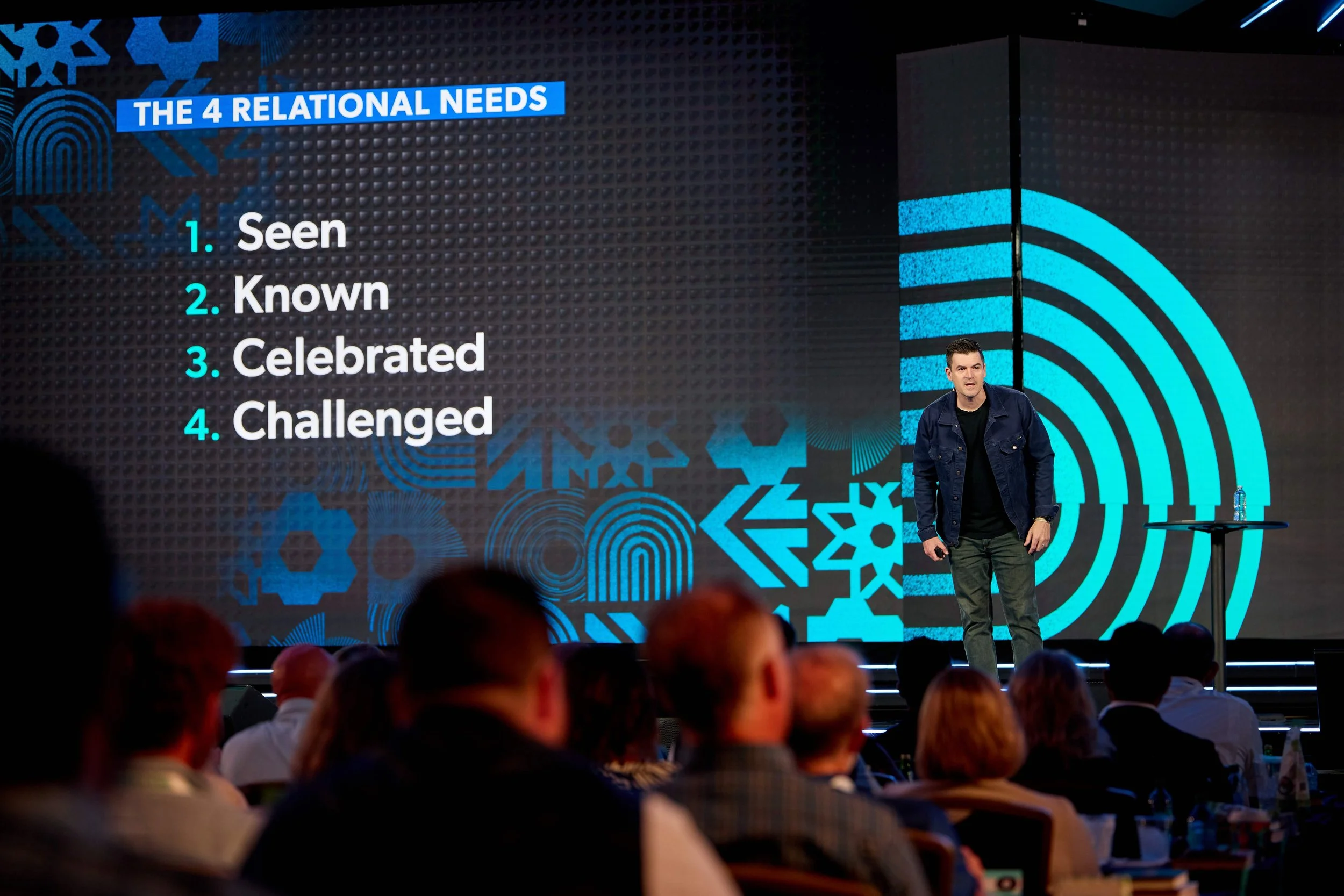 A man is standing on stage with a large screen behind him displaying the title 'The 4 Relational Needs' and a numbered list: 1. Seen, 2. Known, 3. Celebrated, 4. Challenged.' The audience is seated and listening.