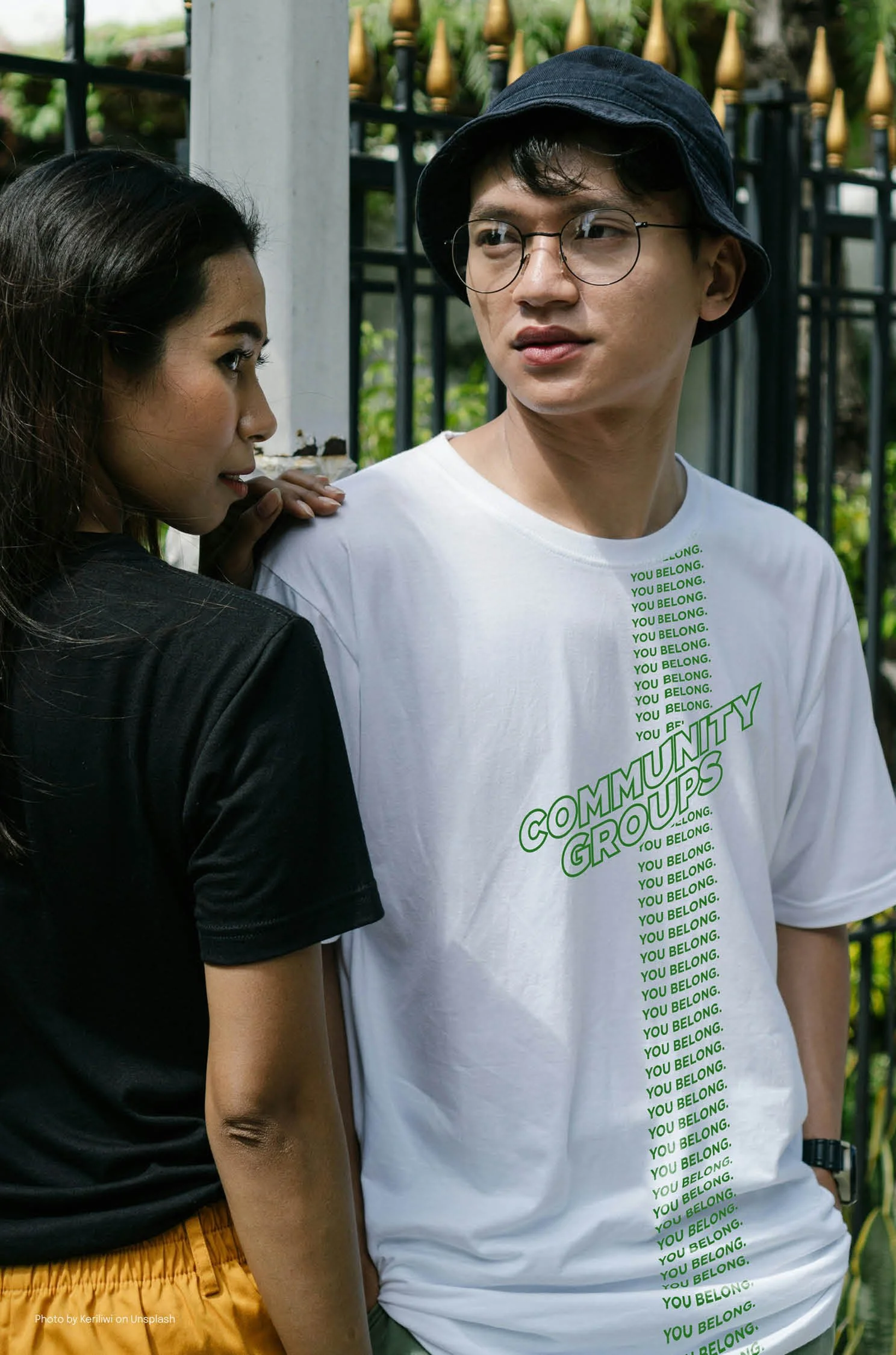 A young man wearing glasses, a navy bucket hat, and a white T-shirt with green text standing outdoors next to a young woman with long dark hair, wearing a black top and yellow skirt. They are near a black metal fence with greenery behind them.
