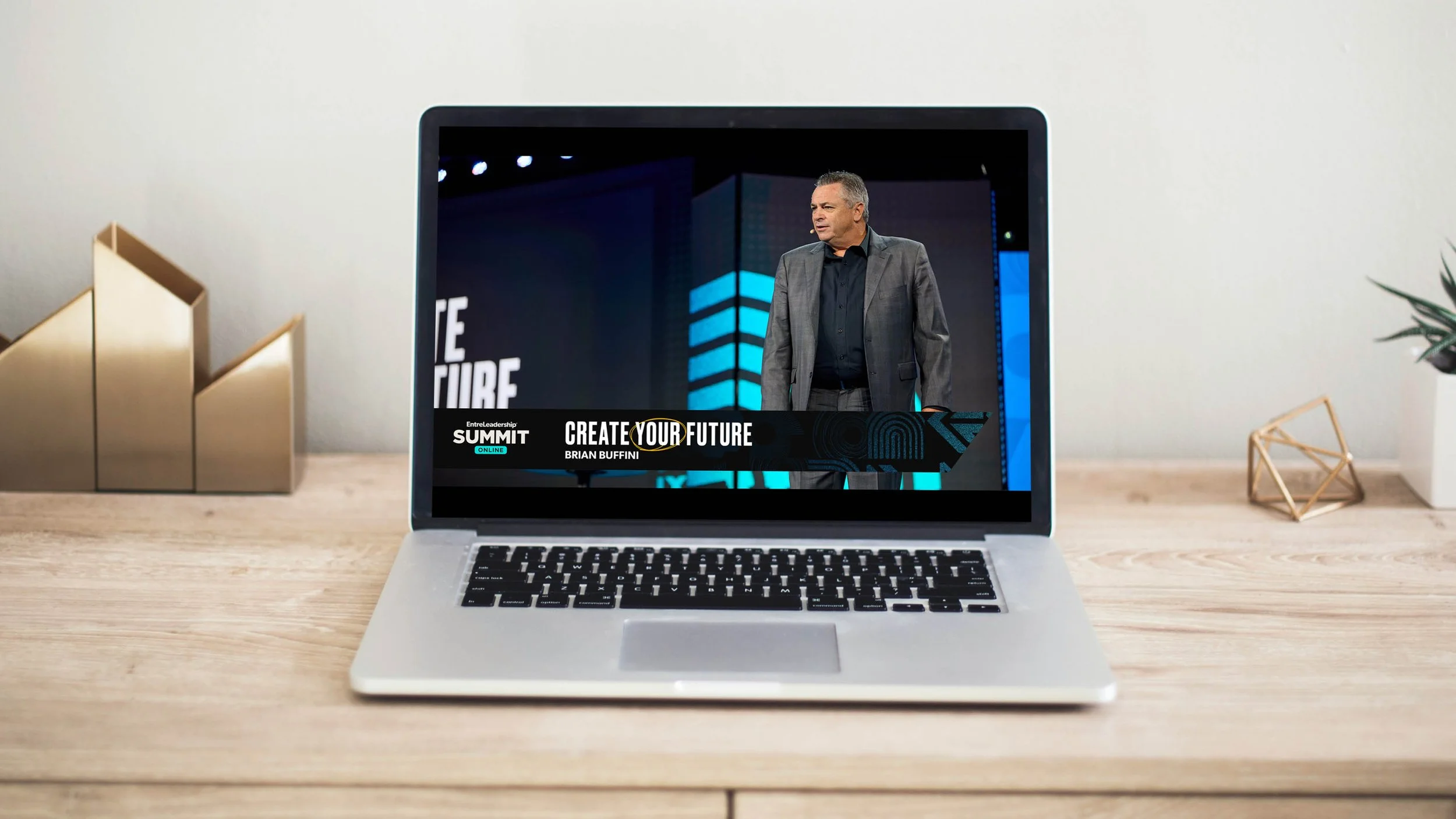 Laptop on a wooden desk displaying a speaker in a suit during a virtual conference with text "CREATE YOUR FUTURE" and "Entreleadership Summit Online".
