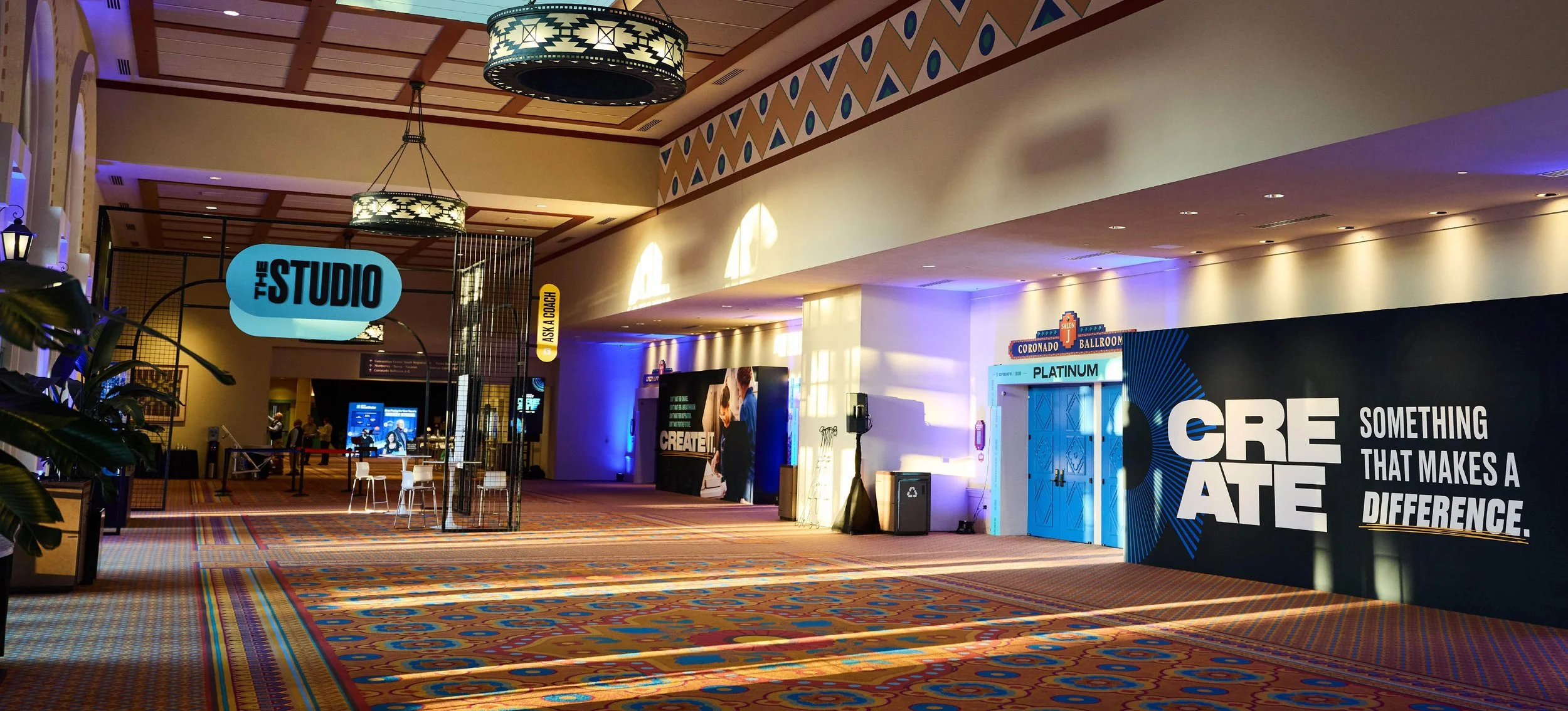 Interior view of a conference or convention center with signs for 'The Studio,' 'Ask a Coach,' and 'Create. Something that makes a difference.' The area features patterned carpet, modern lighting, and a few people in the background.