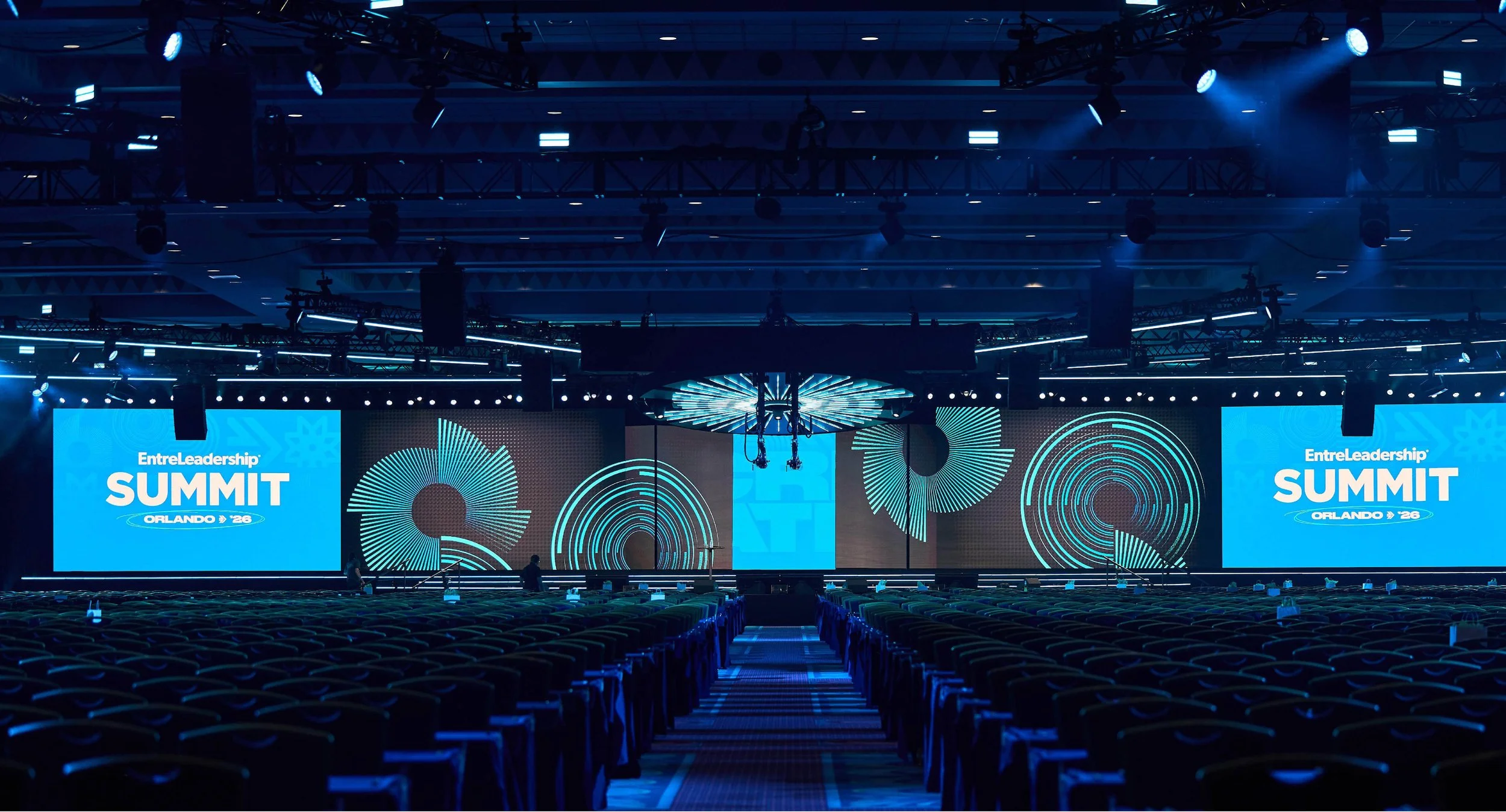 Large conference room with rows of empty chairs facing multiple big screens displaying 'EntreLeadership Summit Orlando 26' in bright blue.
