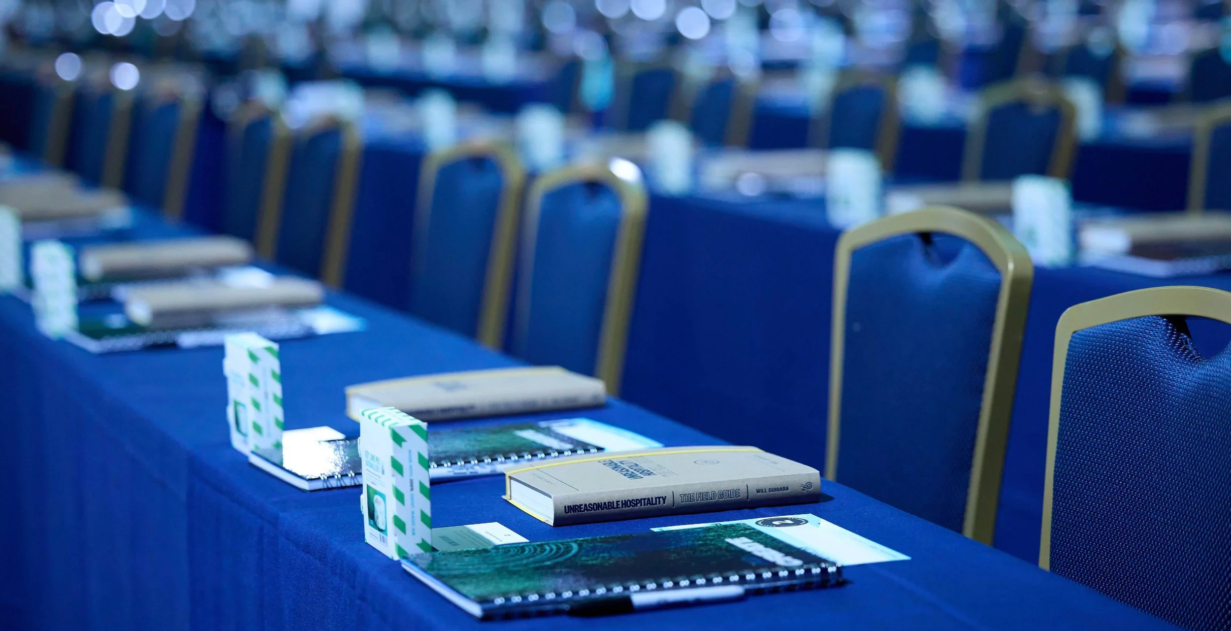 Conference room with rows of blue chairs and tables set up with books, notepads, and pens.