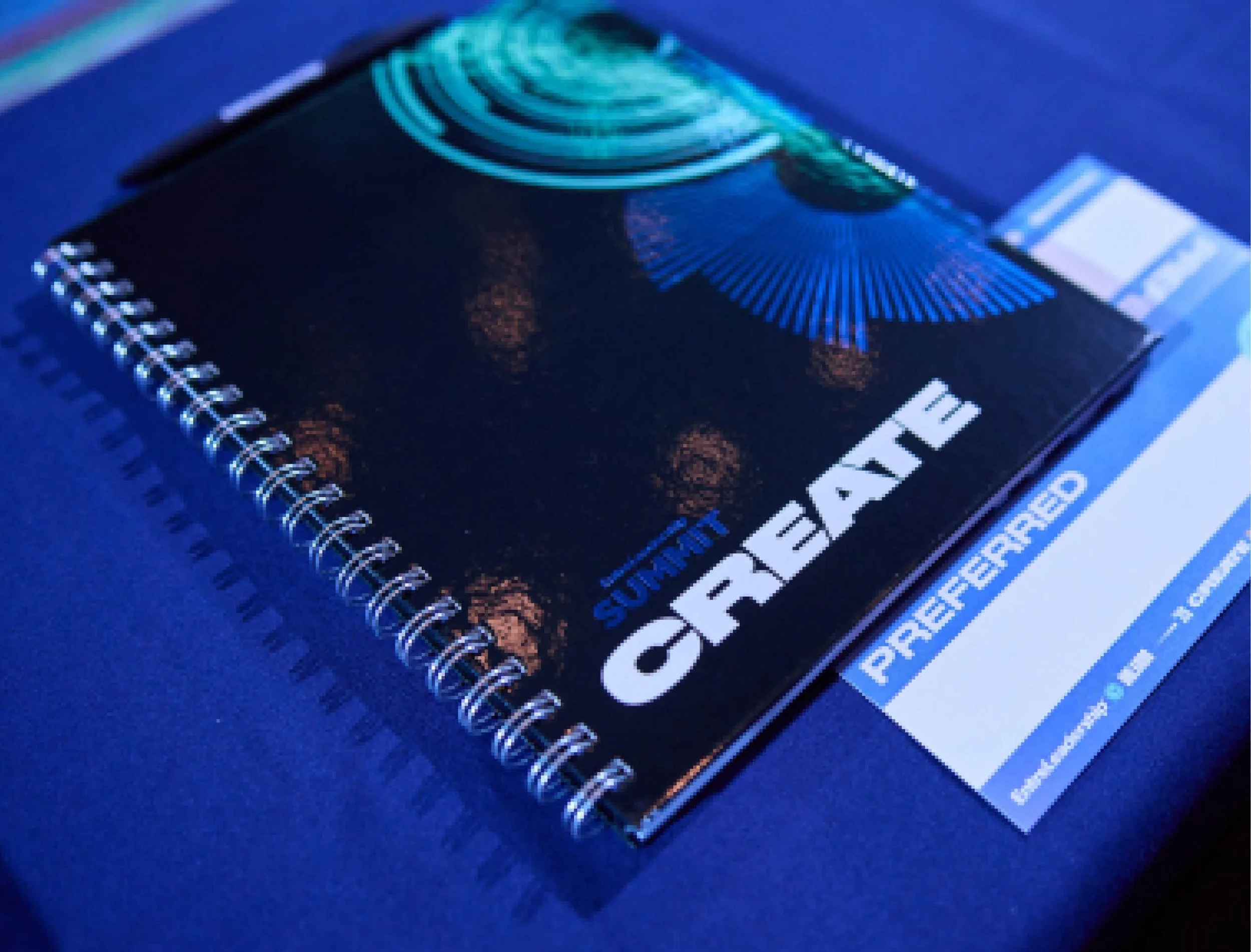 Close-up of a spiral-bound workbook with a printed cover featuring a metallic foiled elements from the create theme, placed on a blue surface with additional printed materials partially visible.