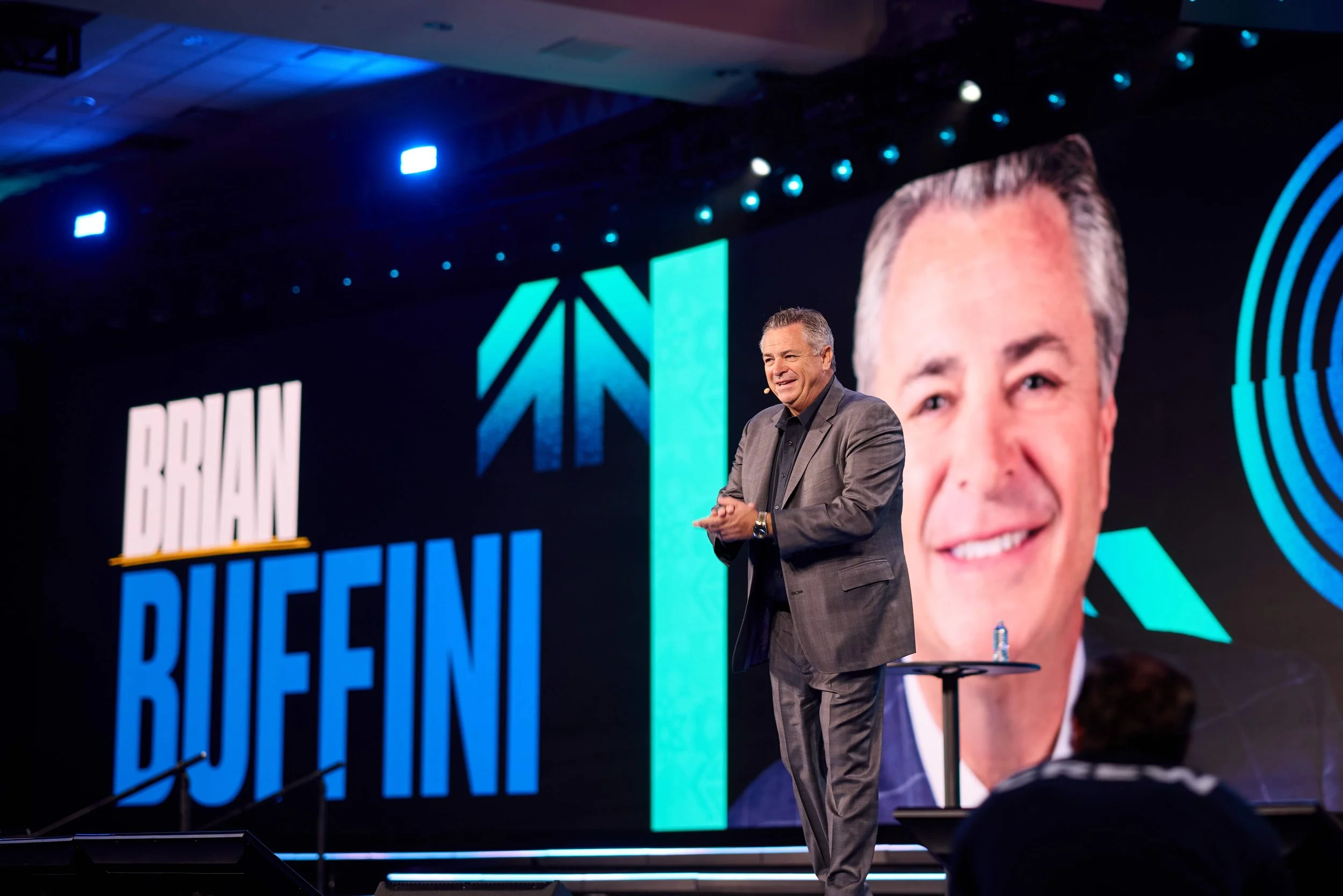 A man in a gray suit speaking on stage with a large screen behind him that shows a large photo of his face and the words 'Brain Buffini' in bold blue and white letters.