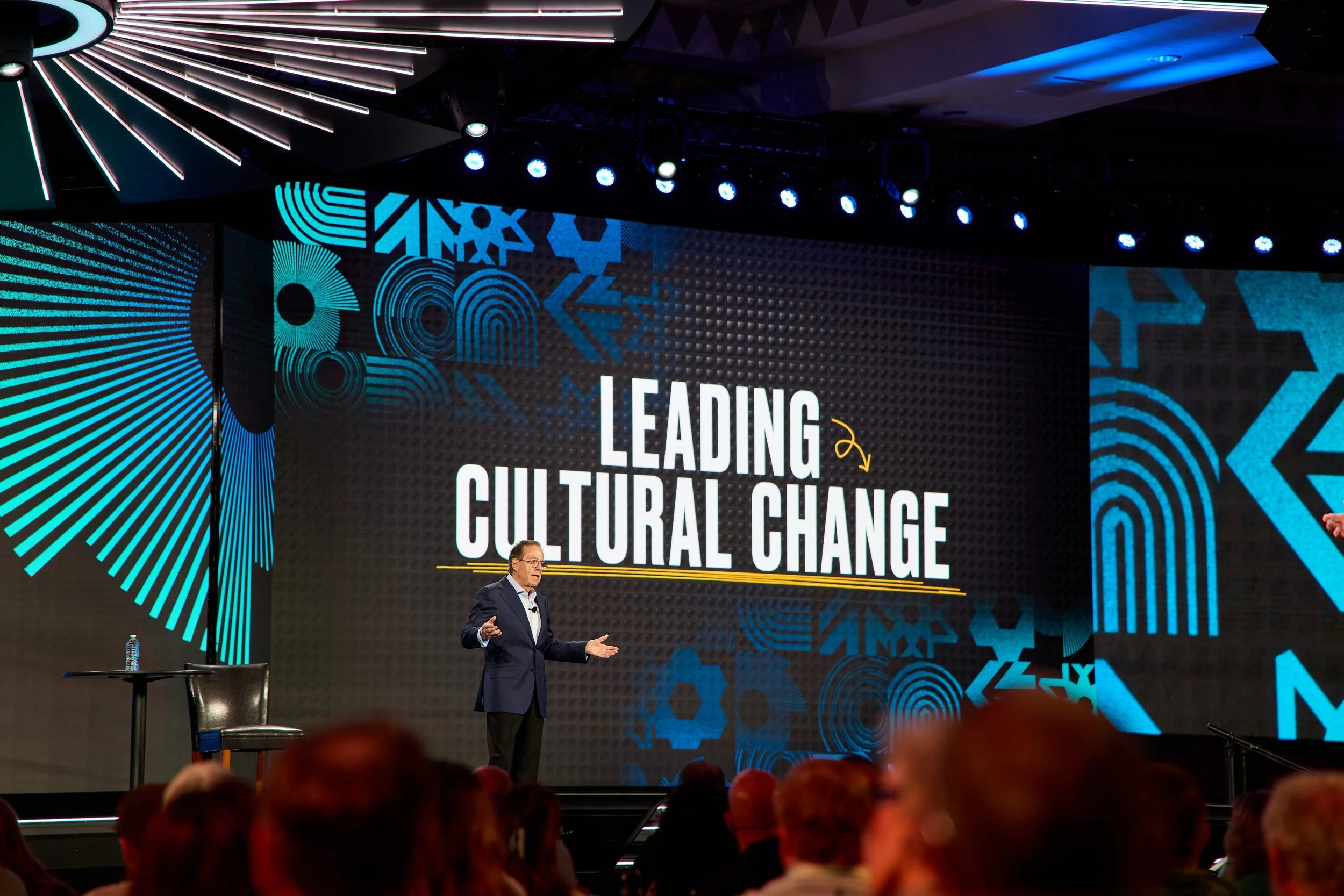 A man in a suit speaking at a conference with a large screen behind him that reads 'Leading Cultural Change'. The stage has a modern design with blue and black graphics, and audience members are visible in the foreground.
