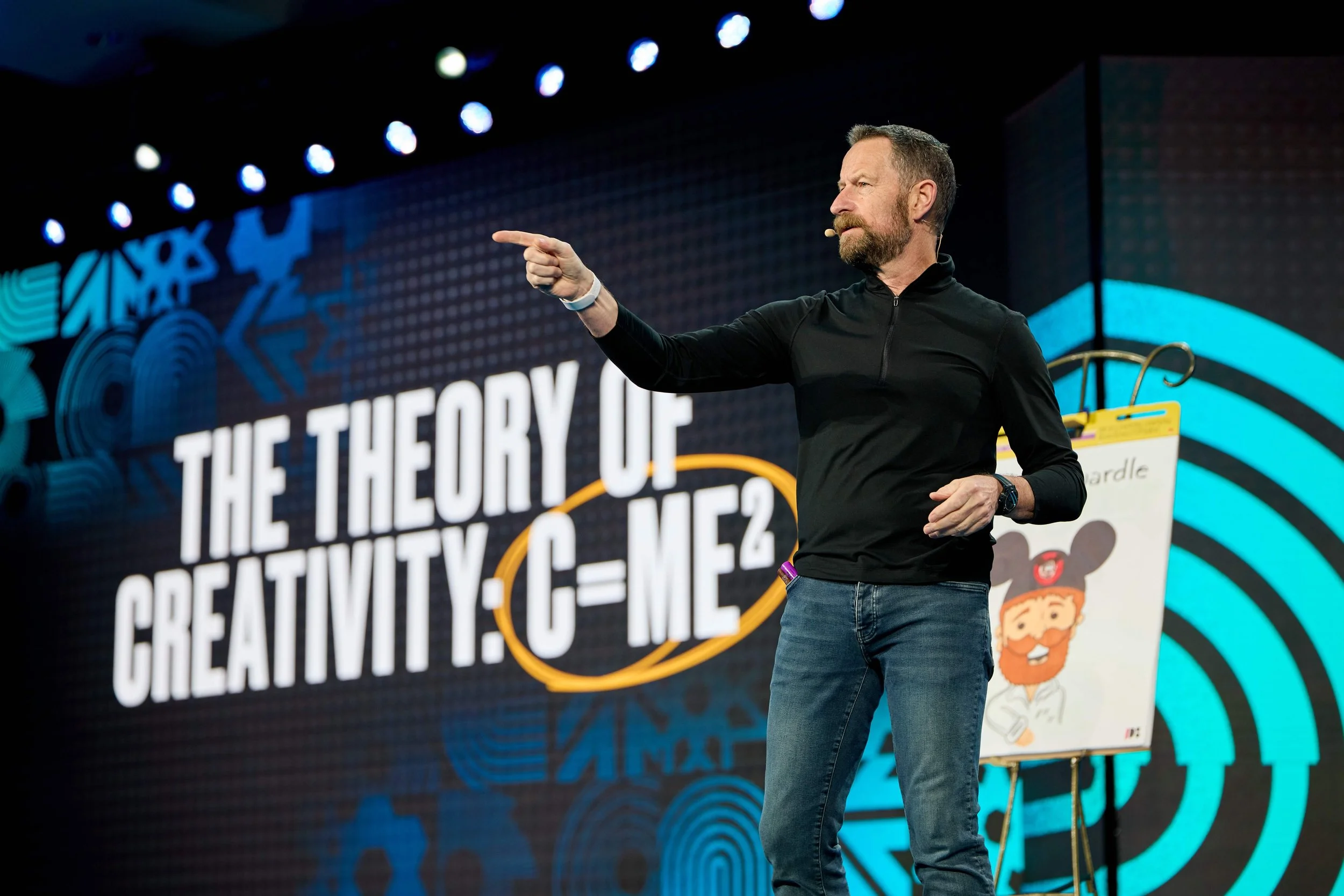 A man with a beard and a black jacket speaking on stage, pointing to his right, with a background displaying the title of his talk, and an easel with a sketch on it.