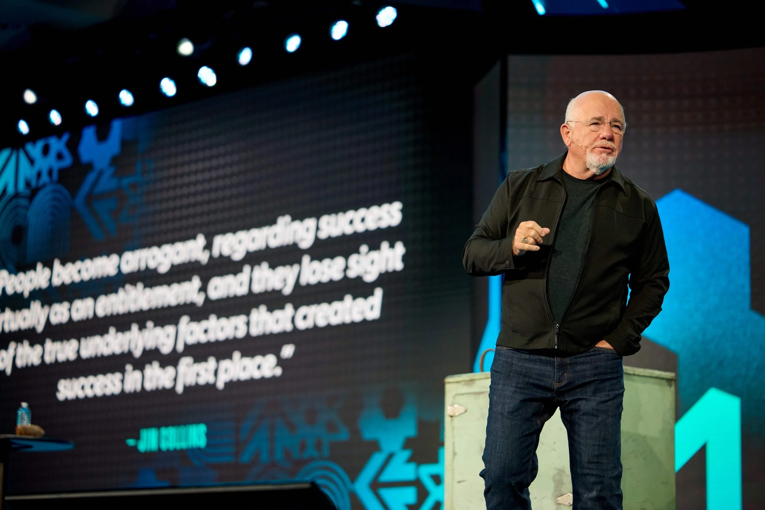A man with a bald head and white beard, wearing glasses, a black jacket, and jeans, stands on stage giving a presentation. Behind him is a large screen with the partially visible text about controlling success and the importance of understanding fact