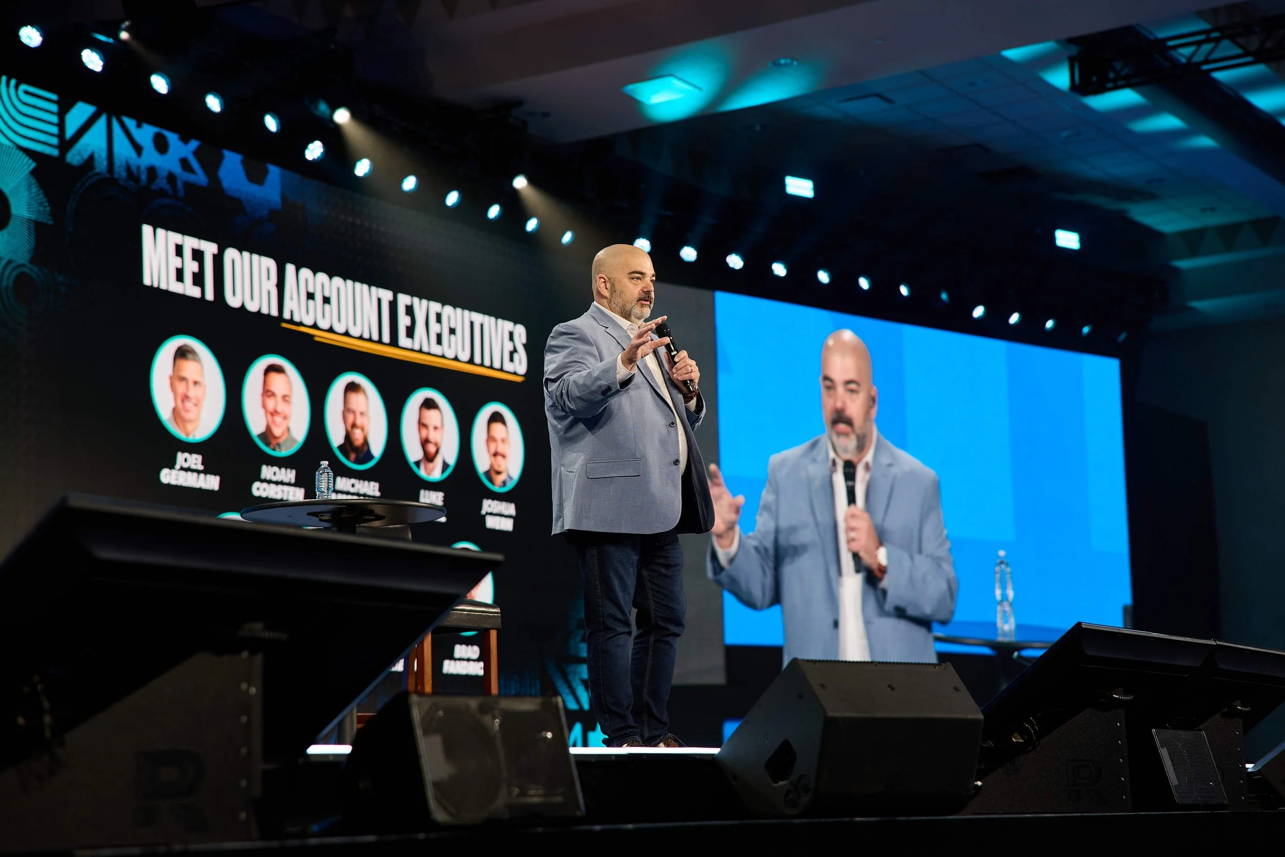 A man in a gray blazer giving a presentation on a stage at a conference. Behind him, a large screen displays the text 'Meet Our Account Executives' and photos of five individuals. The stage is lit with multiple blue and white spotlights, and the back
