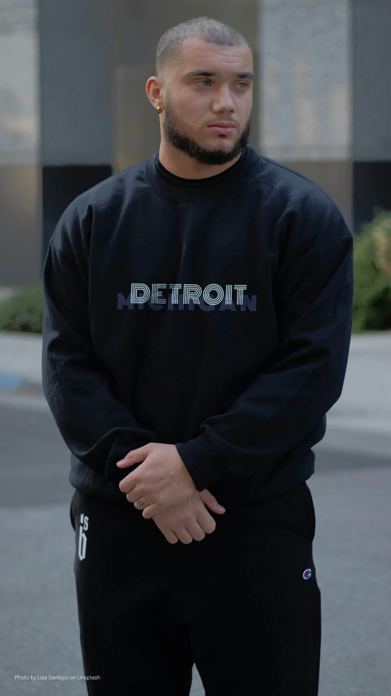 A young man with a buzz cut, beard, and earrings, wearing a black sweatshirt with 'DETROIT' printed on it, standing outdoors with modern buildings in the background.