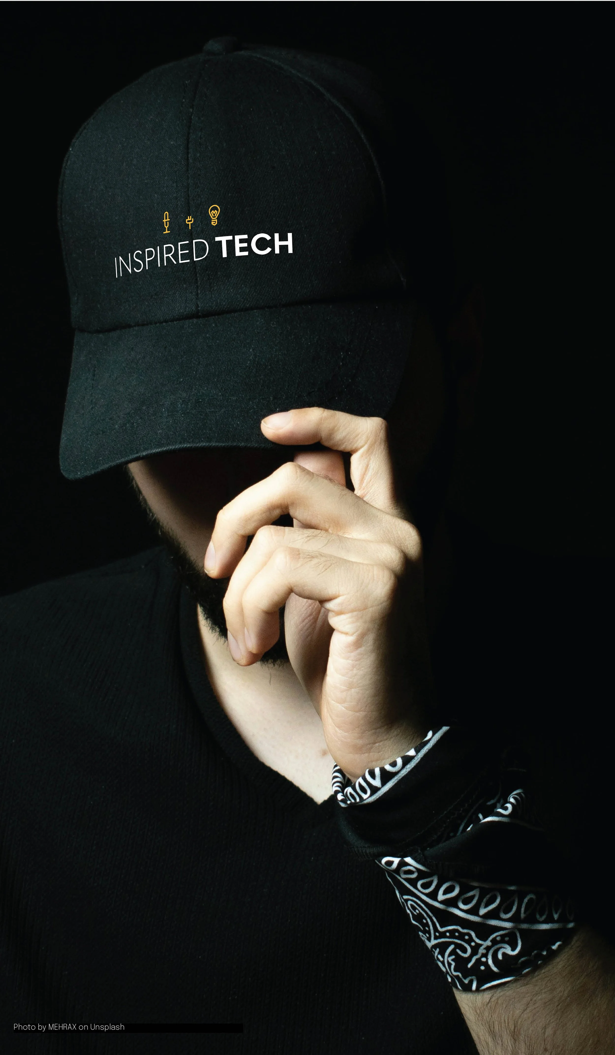 A person wearing a black cap with the words 'INSPIRED TECH' and icons of an electrical plug, a light bulb, microphone. The individual is holding the brim of the cap with one hand, partially obscuring their face. 
